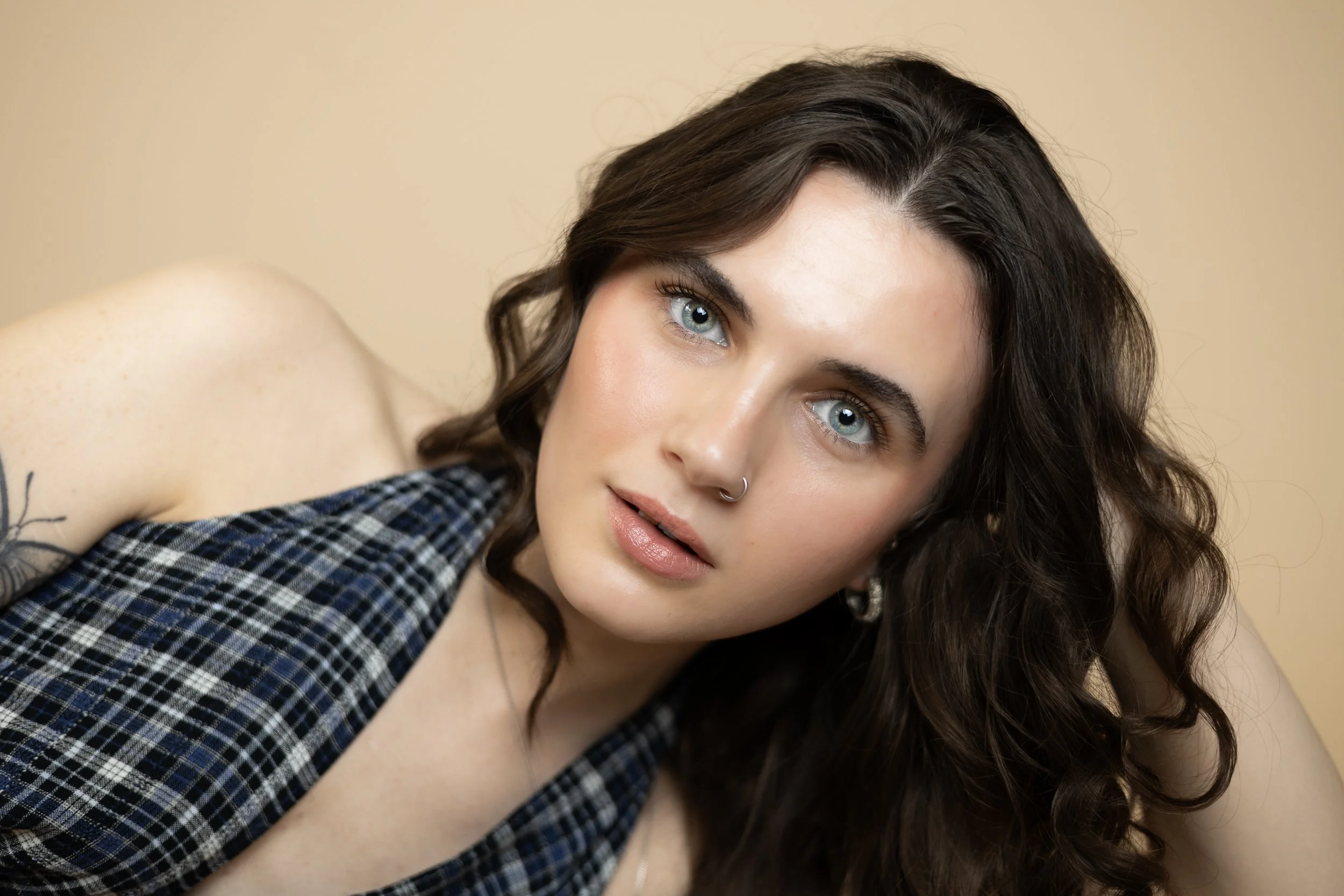 Close-up of a woman with curly brown hair, blue eyes, and a nose ring, wearing a black and white plaid shirt, posed against a beige background.