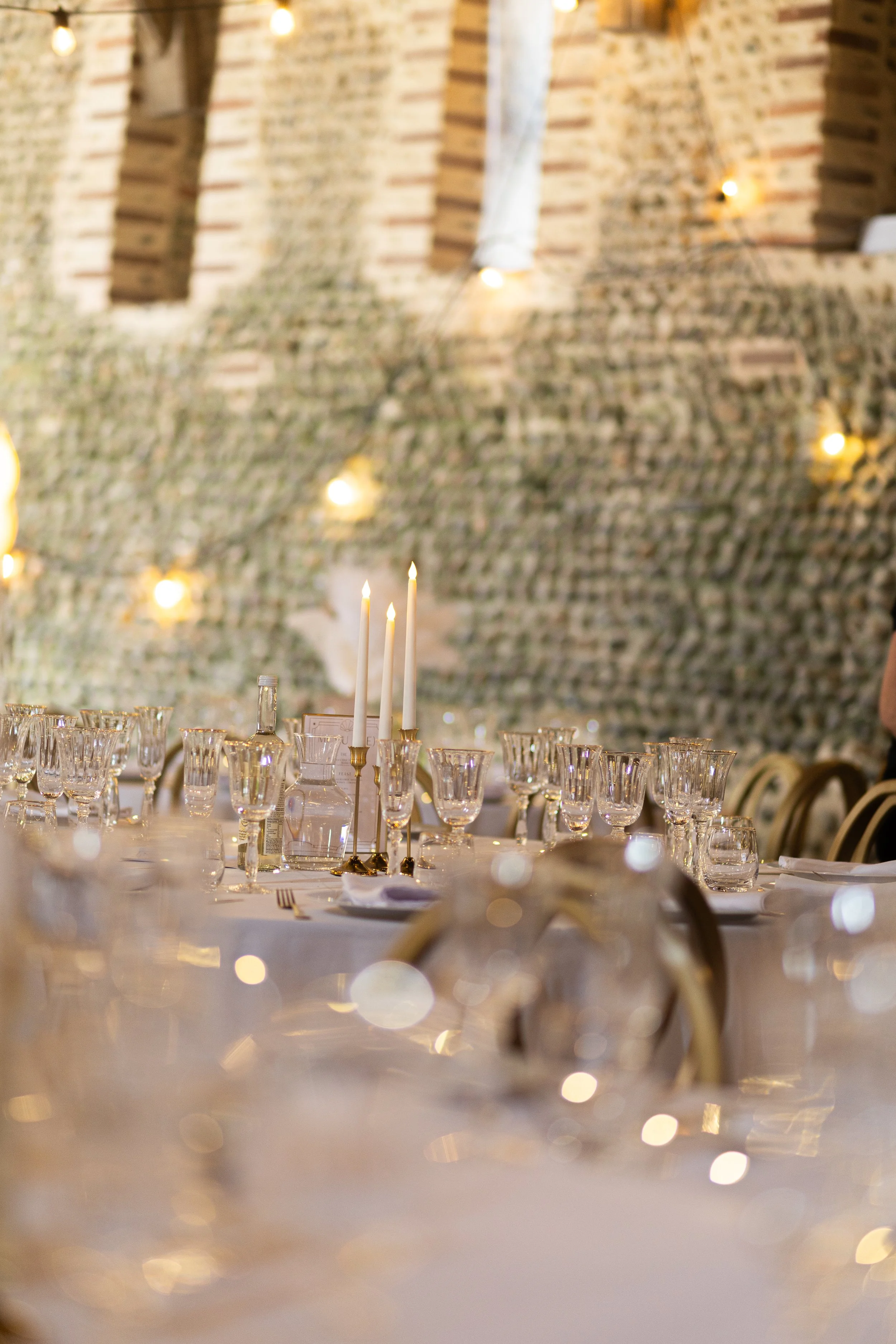 Elegant table setting with glassware, candles, and gold accents in a brick-walled venue decorated with string lights.