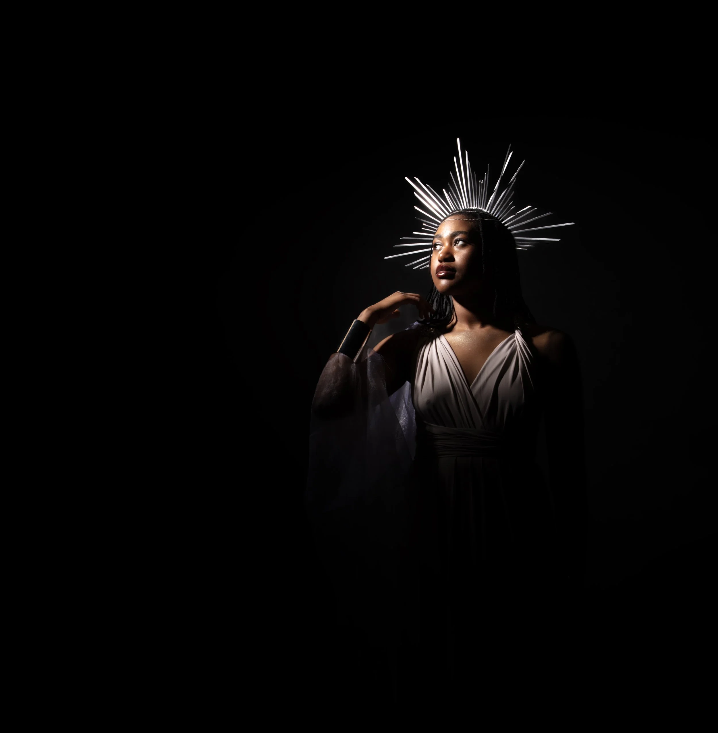 A woman with dark hair and makeup, wearing a white dress, is captured with a black background. She has a headpiece resembling a radiant crown made of metallic rods that fans out from her head.