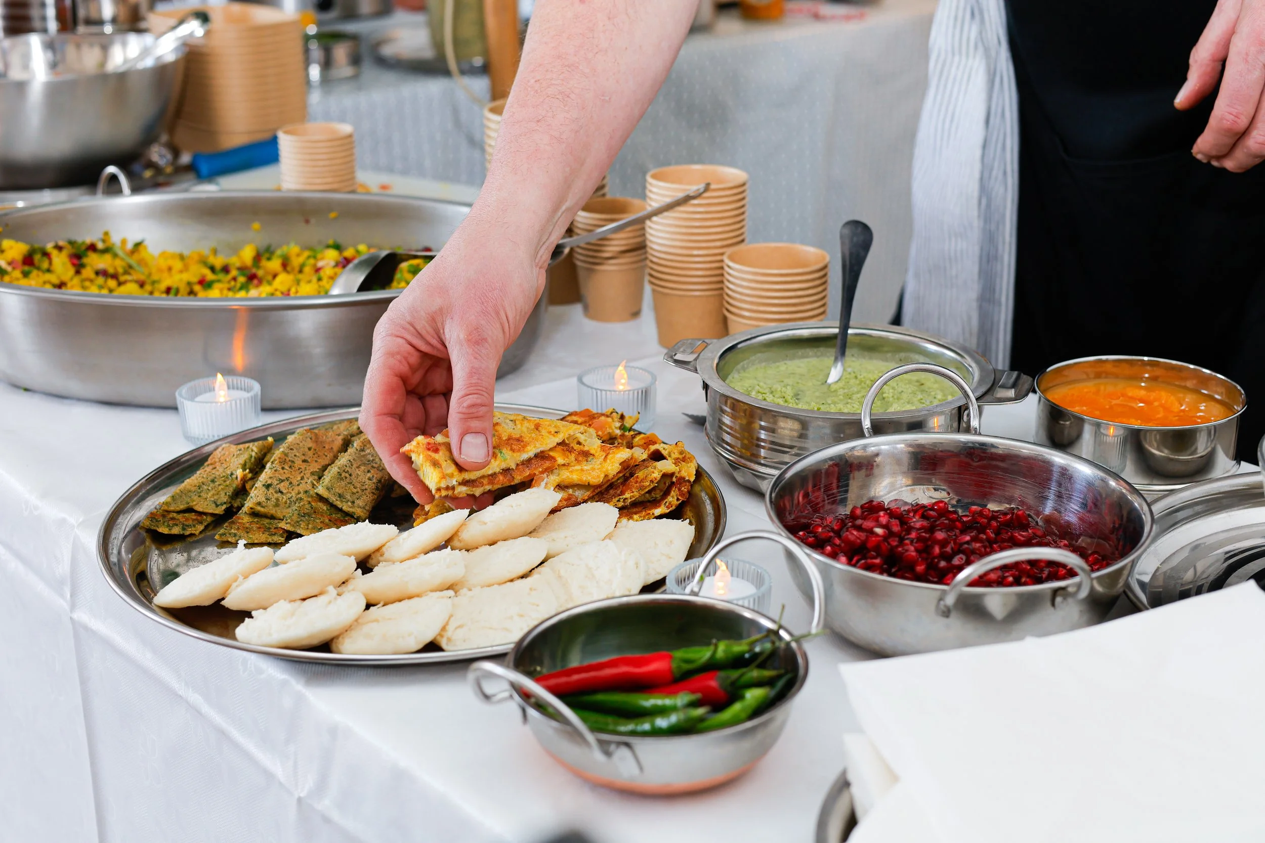 A person serving food at a buffet with various dishes including sliced bread, green chutney, pomegranate seeds, and pastries on a white tablecloth.