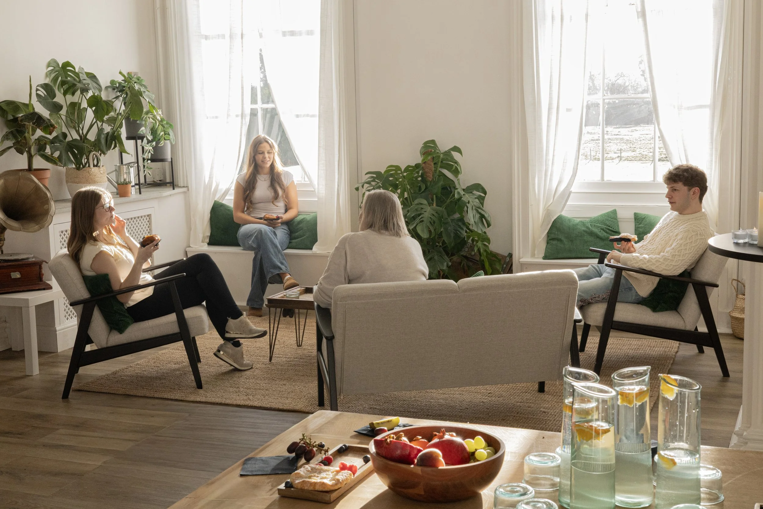 A group of people sitting in a bright living room, enjoying snacks and conversation. There are large windows with white curtains, green cushions, and various potted plants, with a table in the foreground holding a fruit bowl and glasses of drinks.