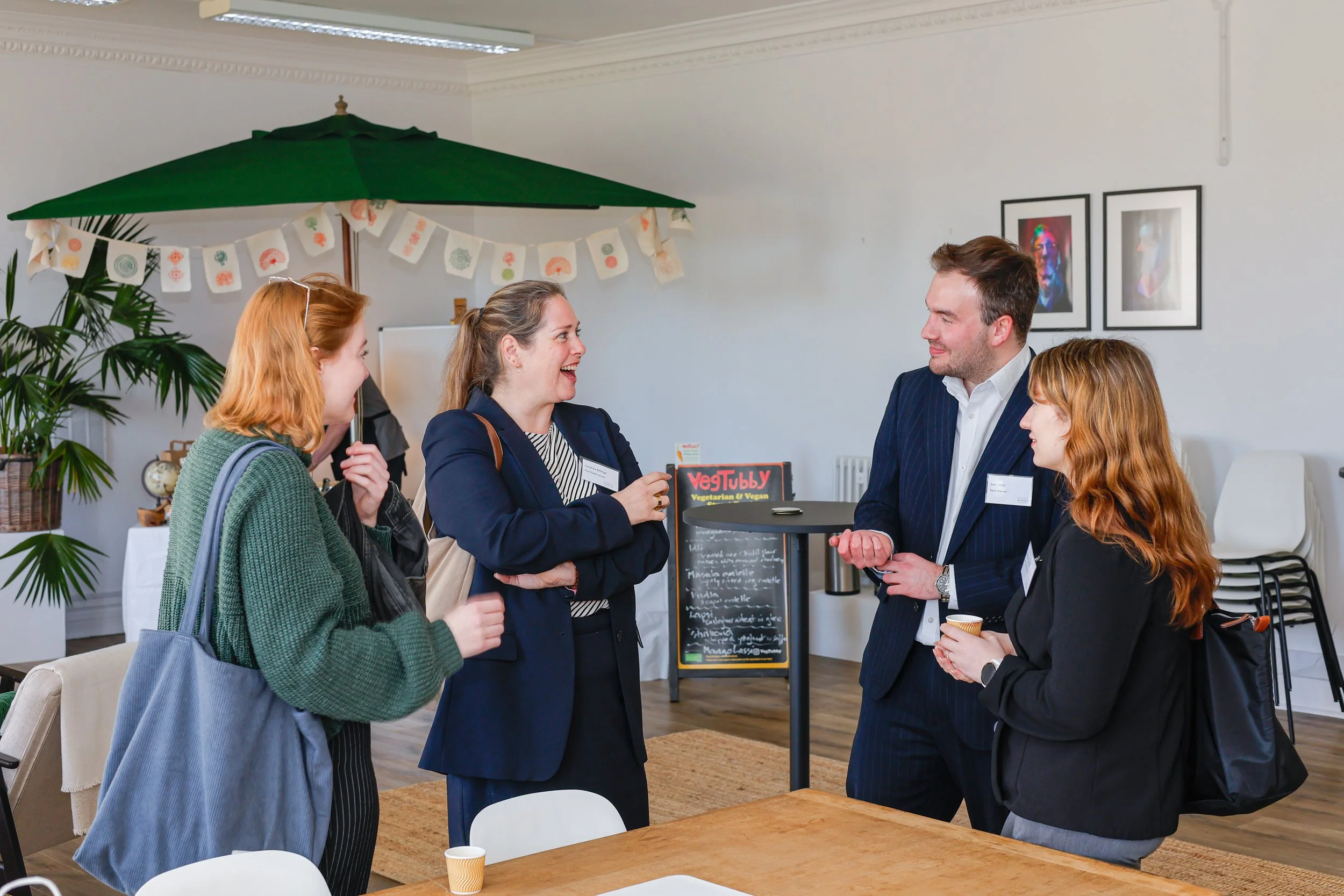 Four women and one man are standing and talking in a room with white walls, framed artwork, and a large green umbrella. They are smiling and engaged in conversation. Some are holding cups, and there is a small chalkboard menu in the background.