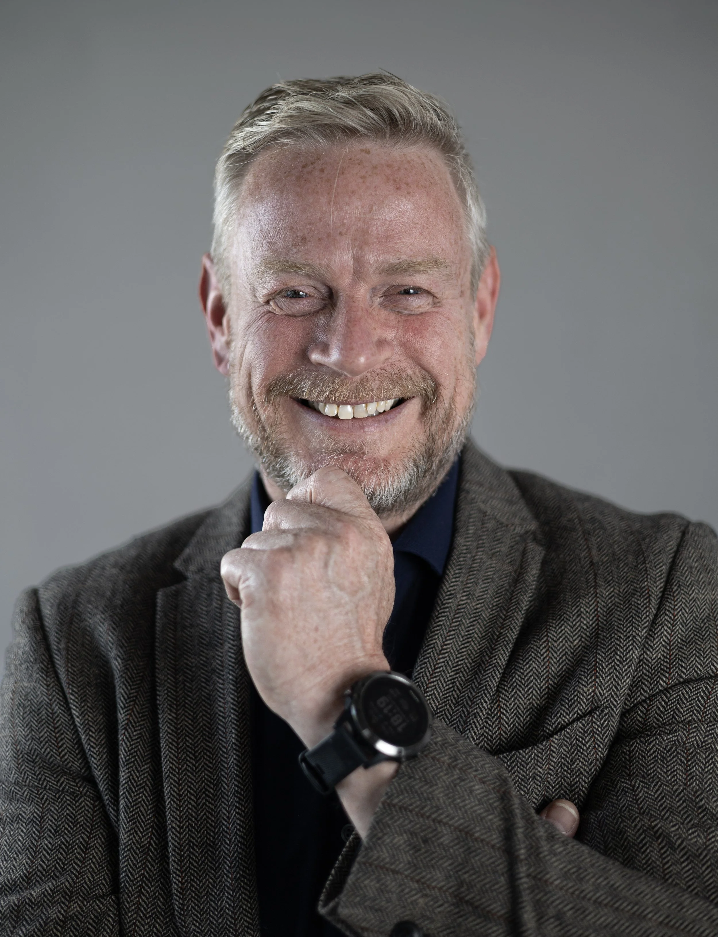 A smiling middle-aged man with gray hair, a beard, and blue eyes, wearing a dark shirt and a herringbone blazer, is looking directly at the camera with his hand resting on his chin. There is a gray background behind him.