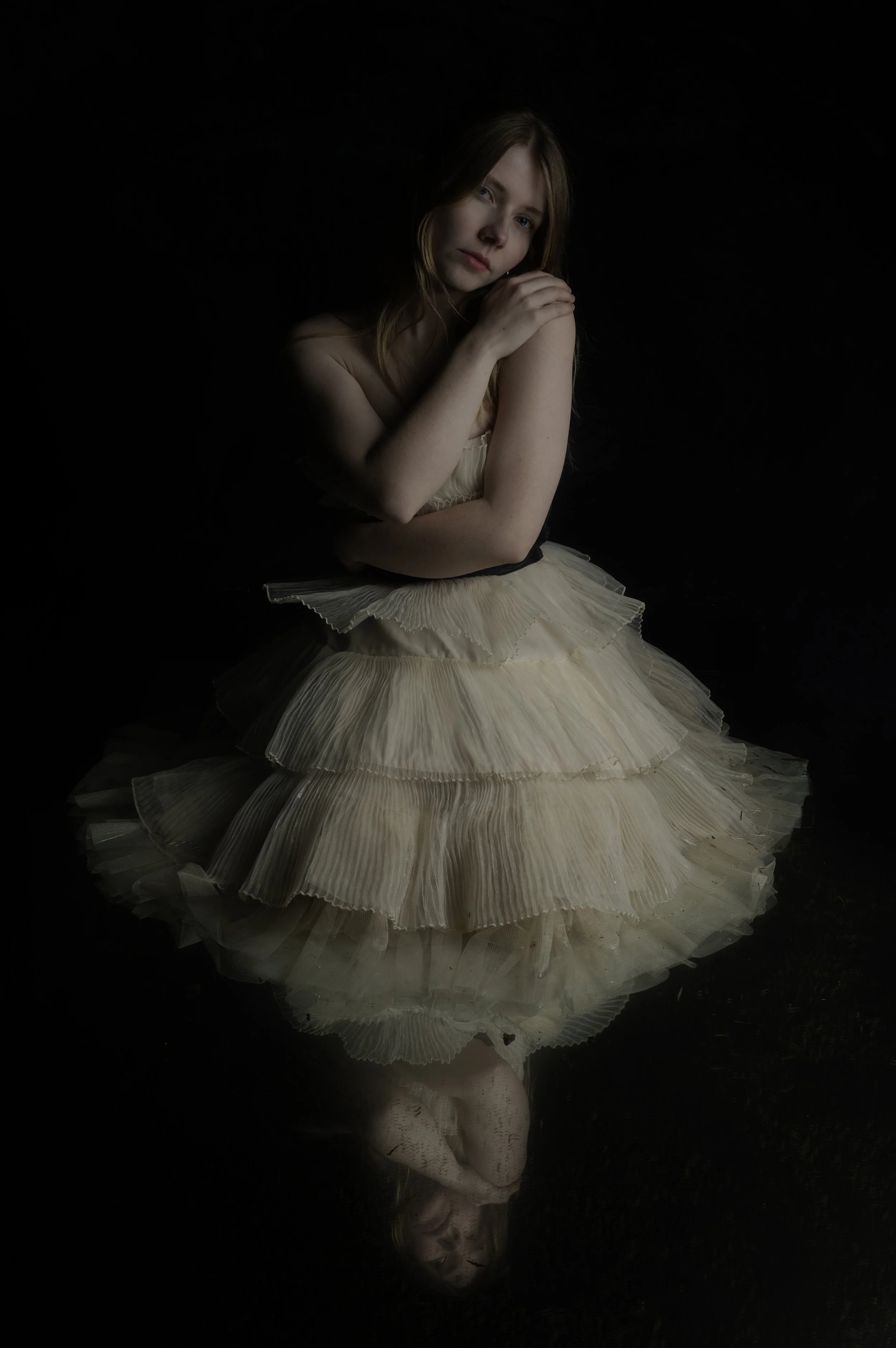 A young woman with long red hair, wearing a cream-colored dress with layered ruffles, sitting against a black background. She has a contemplative expression with one hand resting on her shoulder and her head slightly tilted.