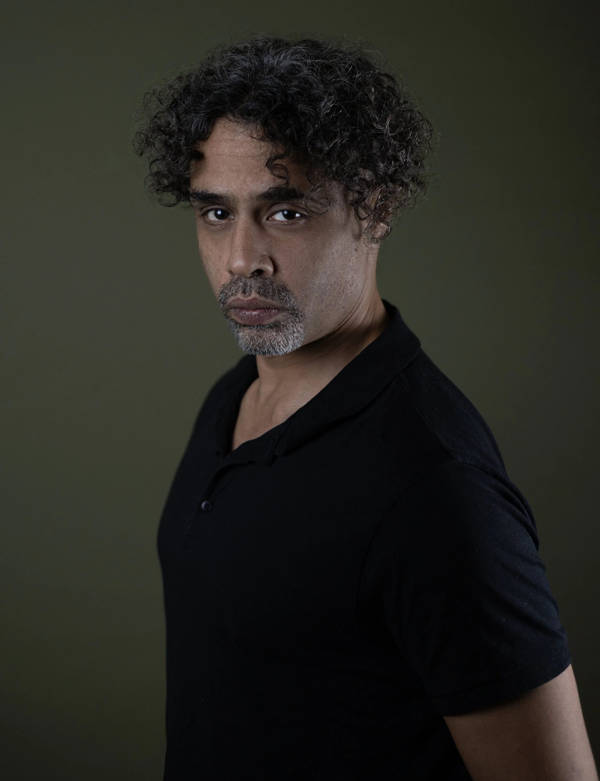 Close-up portrait of a man with curly dark hair, brown eyes, light stubble, and wearing a black shirt against a dark green background.