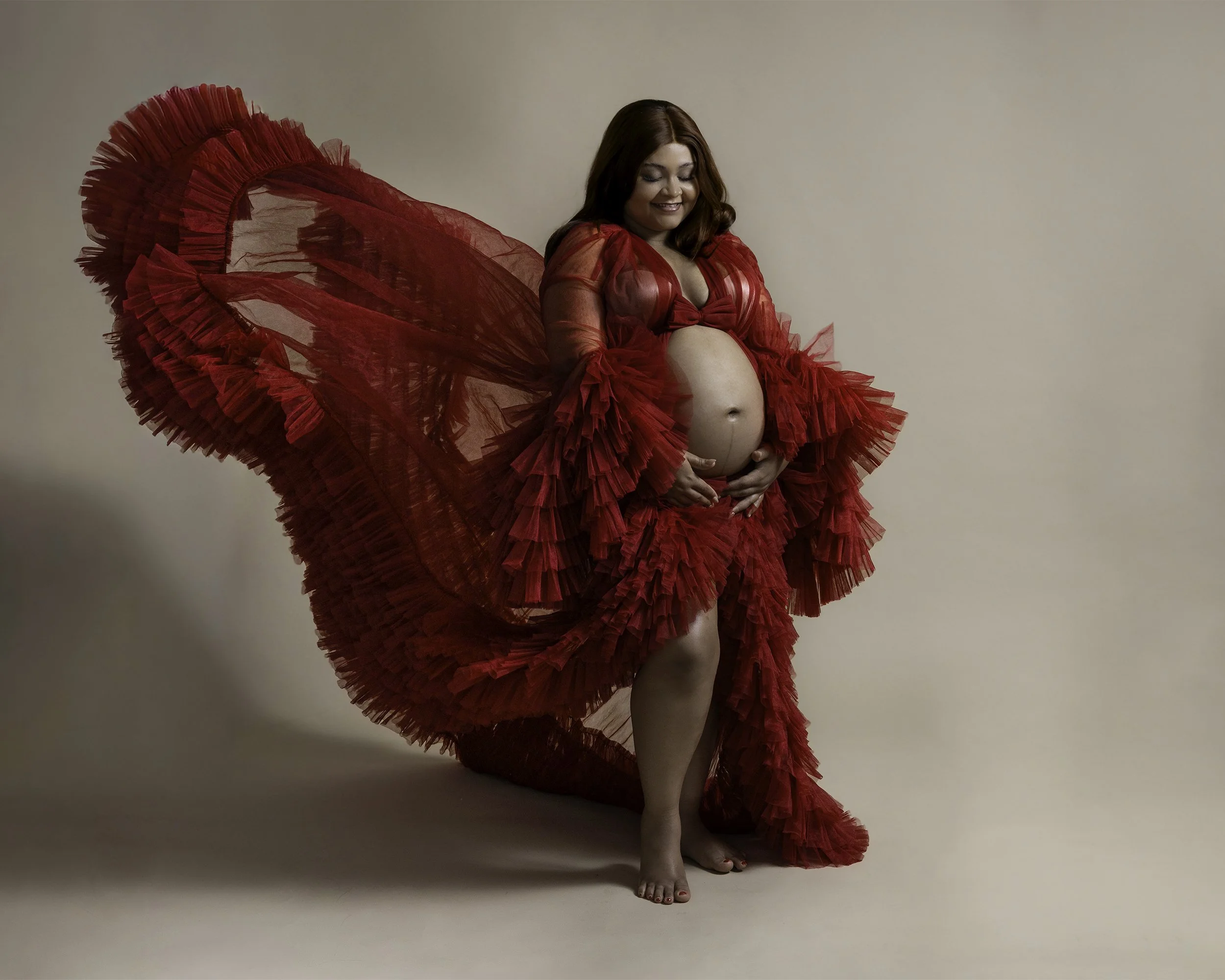 A pregnant woman in a vibrant red, ruffled gown with a large, flowing train, smiling softly, standing against a plain beige background.