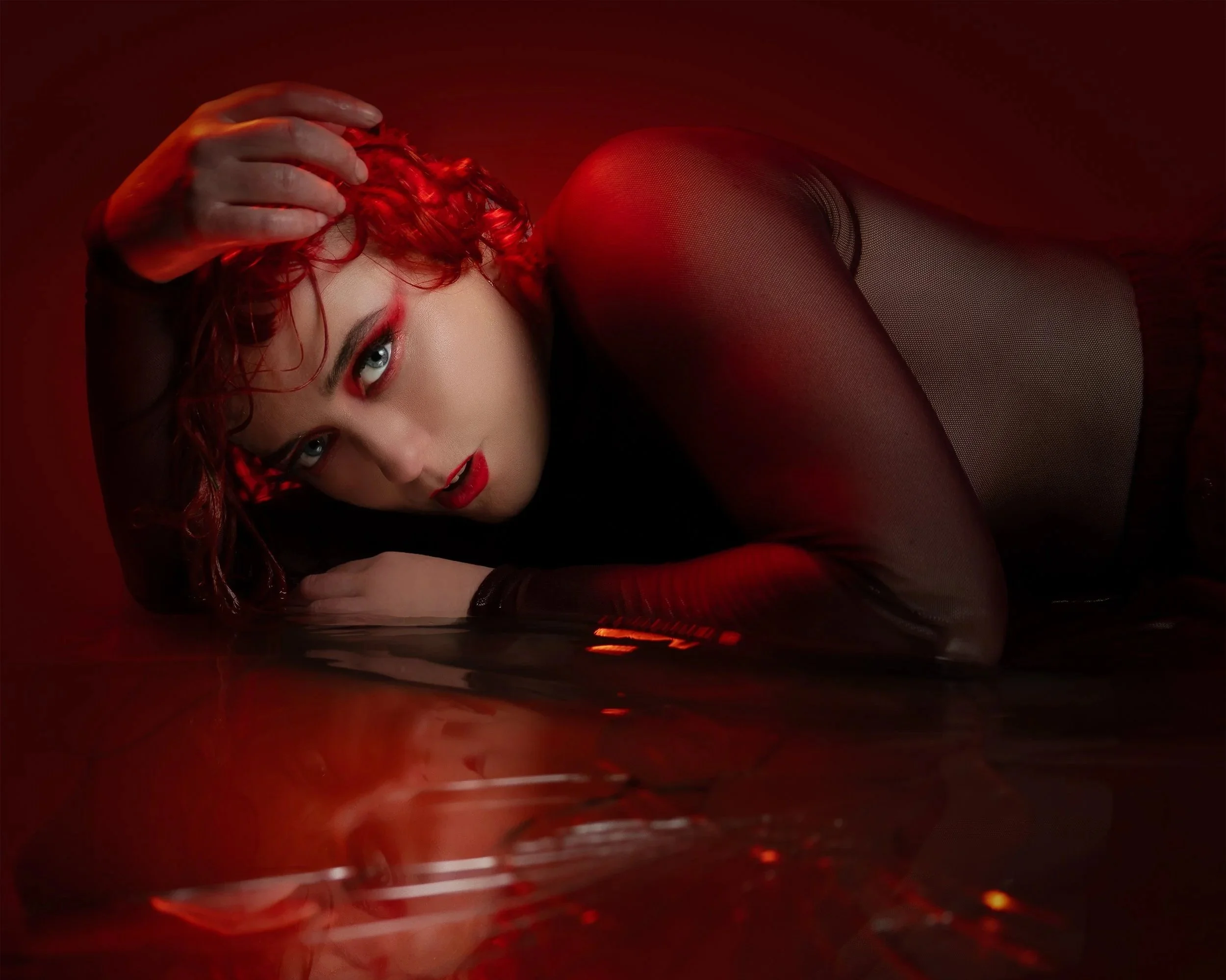 A woman with red curly hair lying on a reflective red surface, wearing sheer black stockings and red lipstick, with a hand on her head and intense eye makeup.