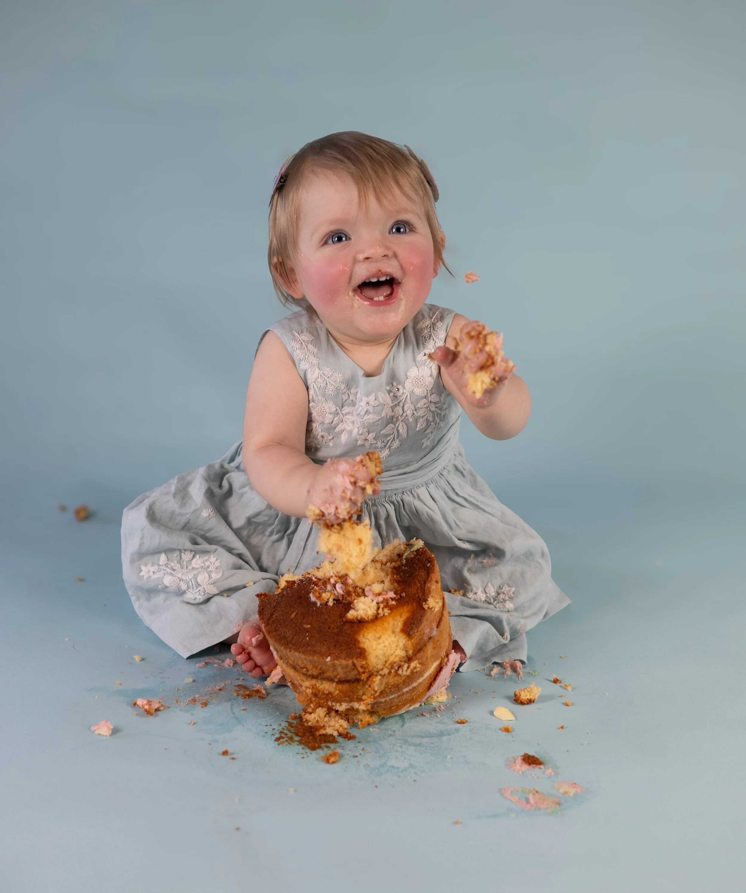 A young girl with light brown hair and blue eyes, wearing a light gray dress with embroidered floral details, sitting on the floor and holding a partially eaten cake with pink frosting and crumbs scattered around.