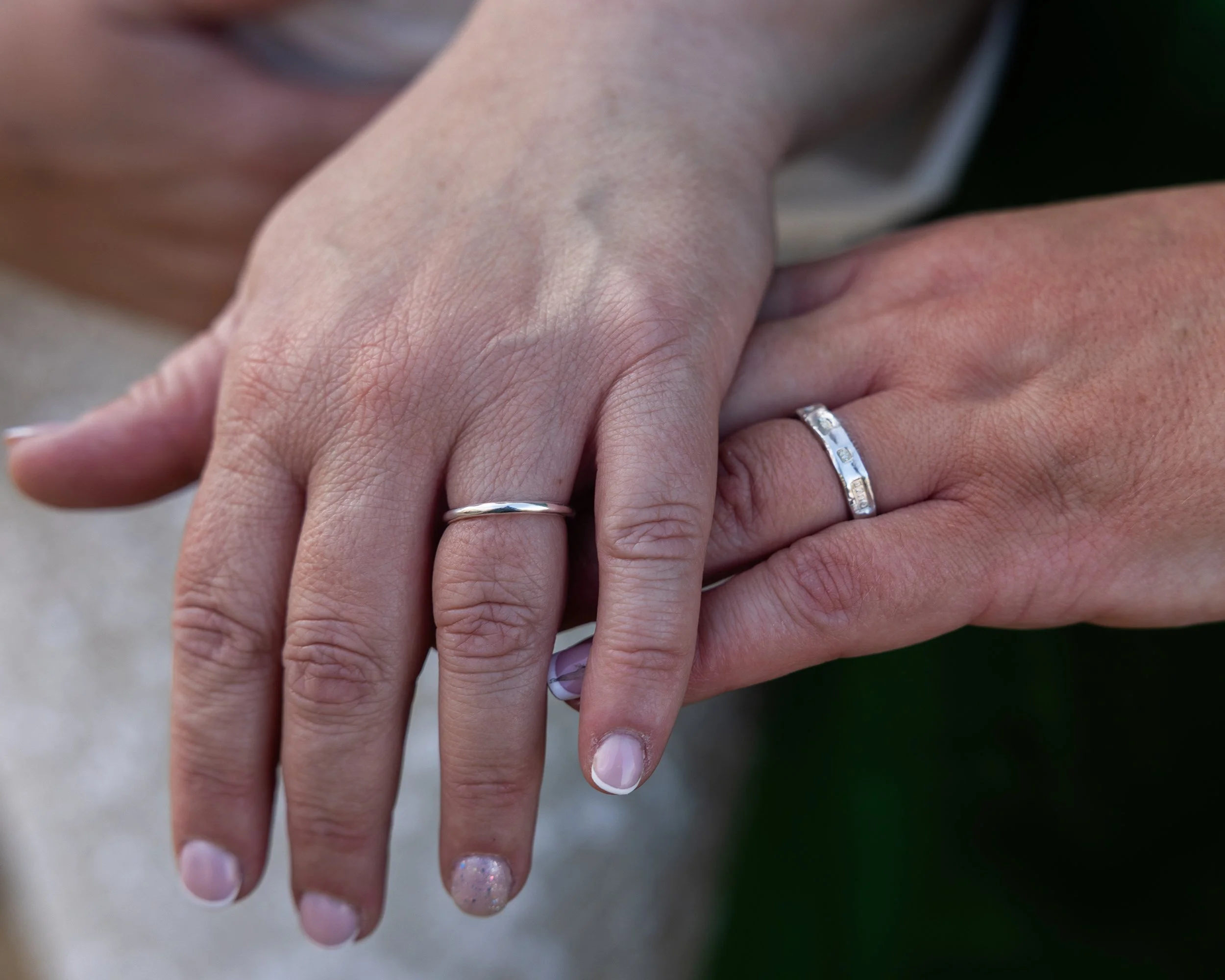 Close-up of hands with rings, one hand wearing a silver band and another hand wearing a ring with a row of small diamonds, indicating a couple holding hands.
