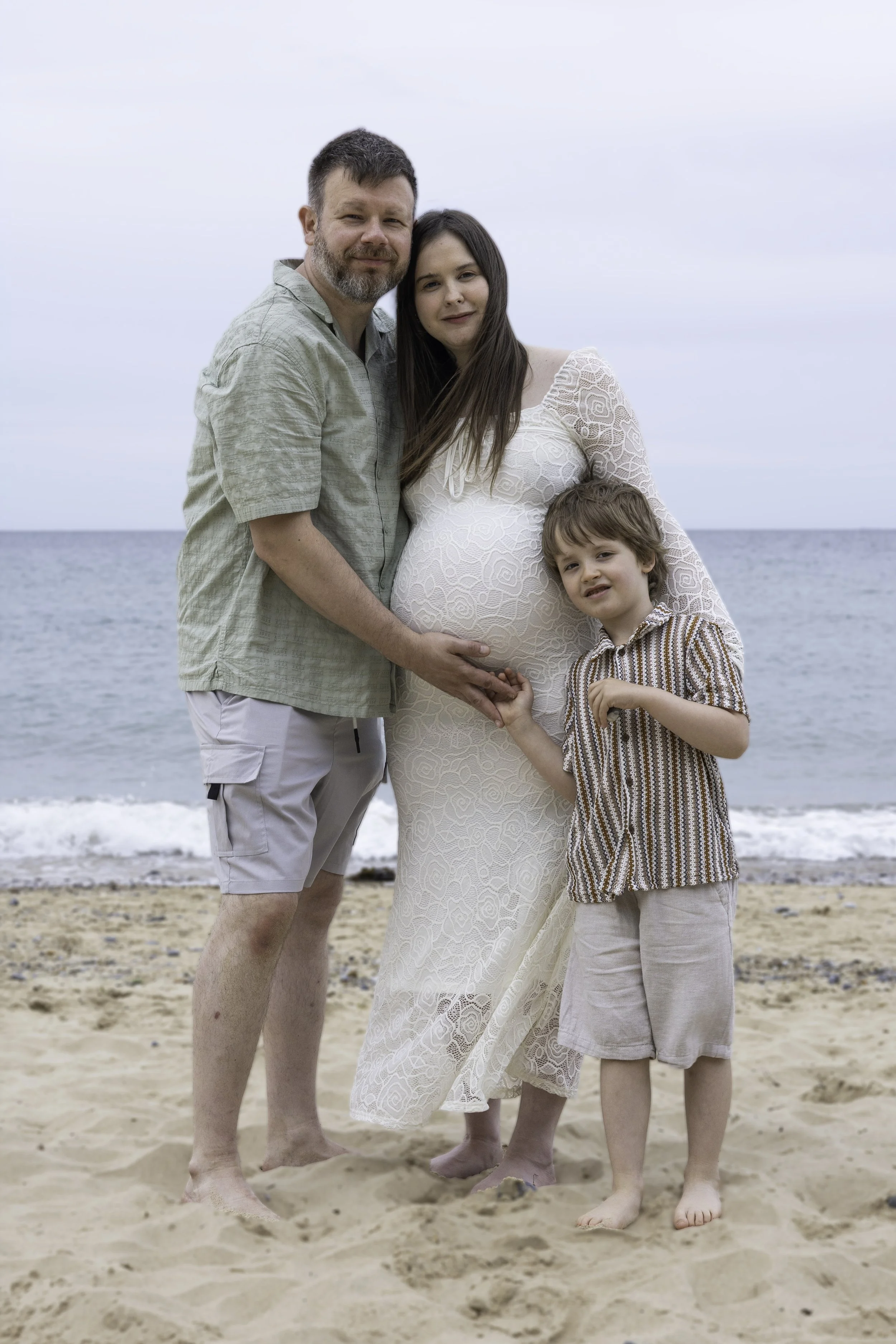 A family of three on the beach, standing close together with the ocean in the background. The pregnant woman is wearing a white lace dress and is holding hands with a young boy. The man, with a beard, is standing next to her with his arm around her s
