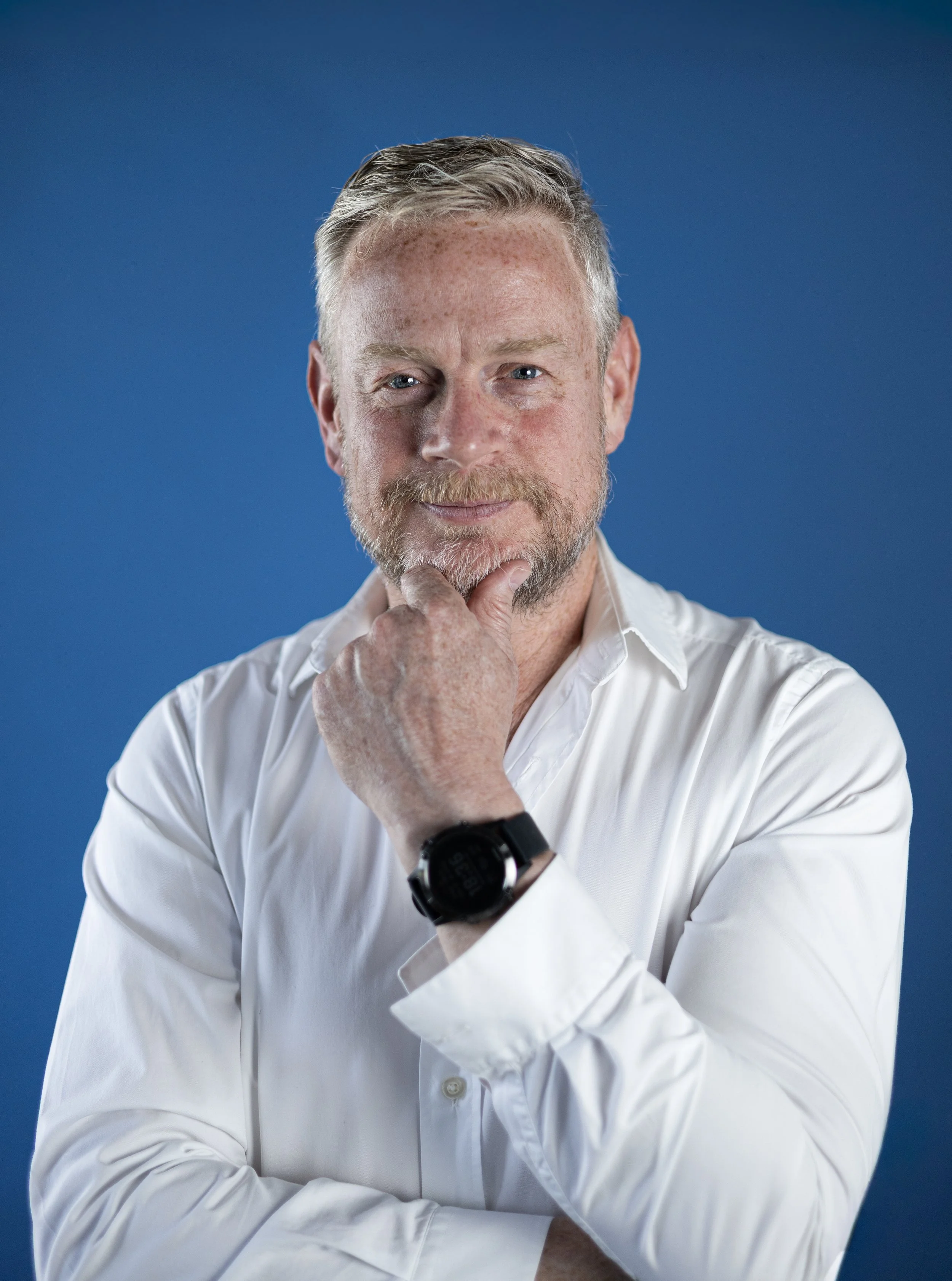 A middle-aged man with gray hair and a beard wearing a white shirt, smiling thoughtfully, against a blue background.