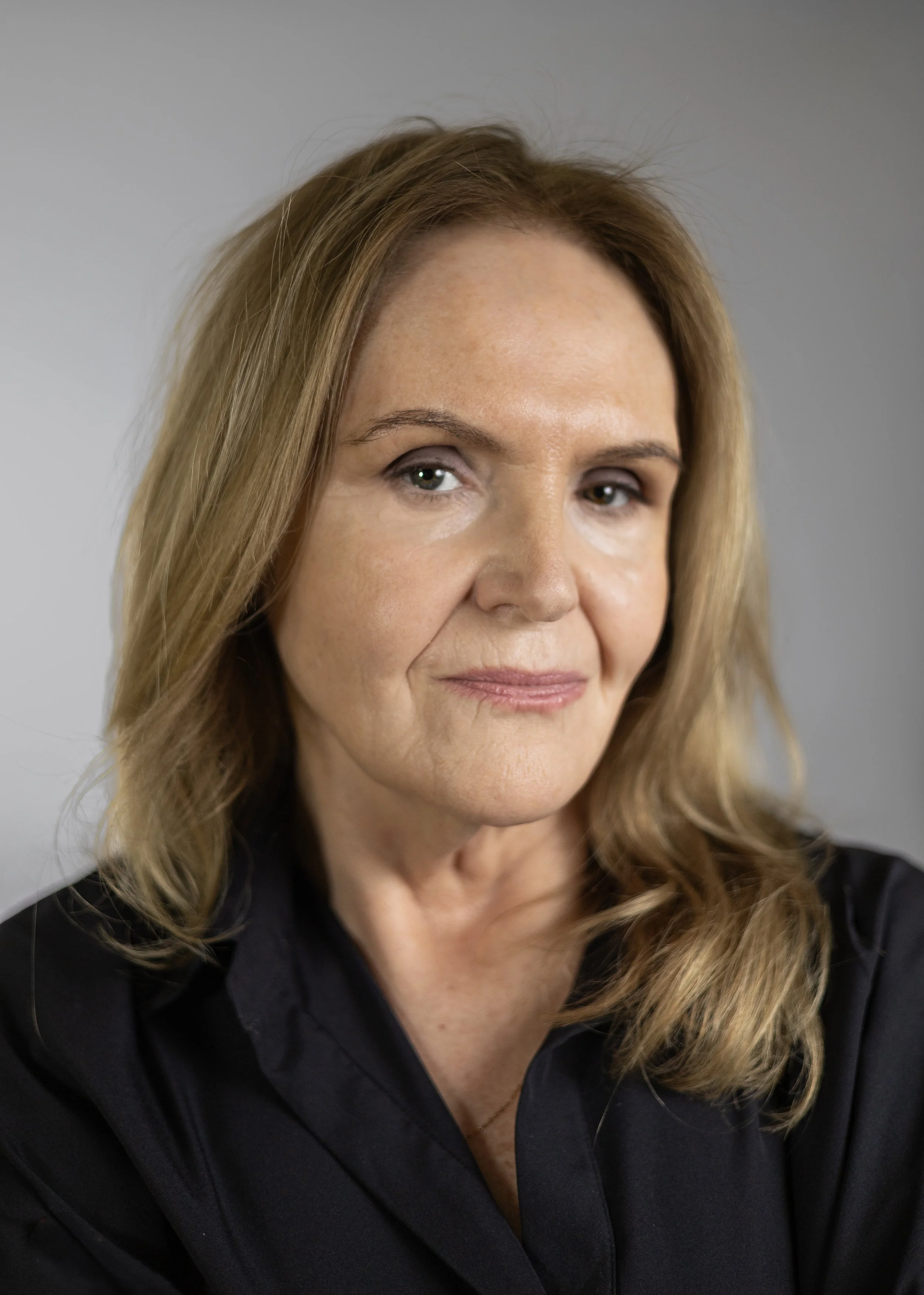 A woman with light skin and wavy blonde hair looking slightly to the side, smiling subtly, wearing a black shirt, in front of a neutral background.