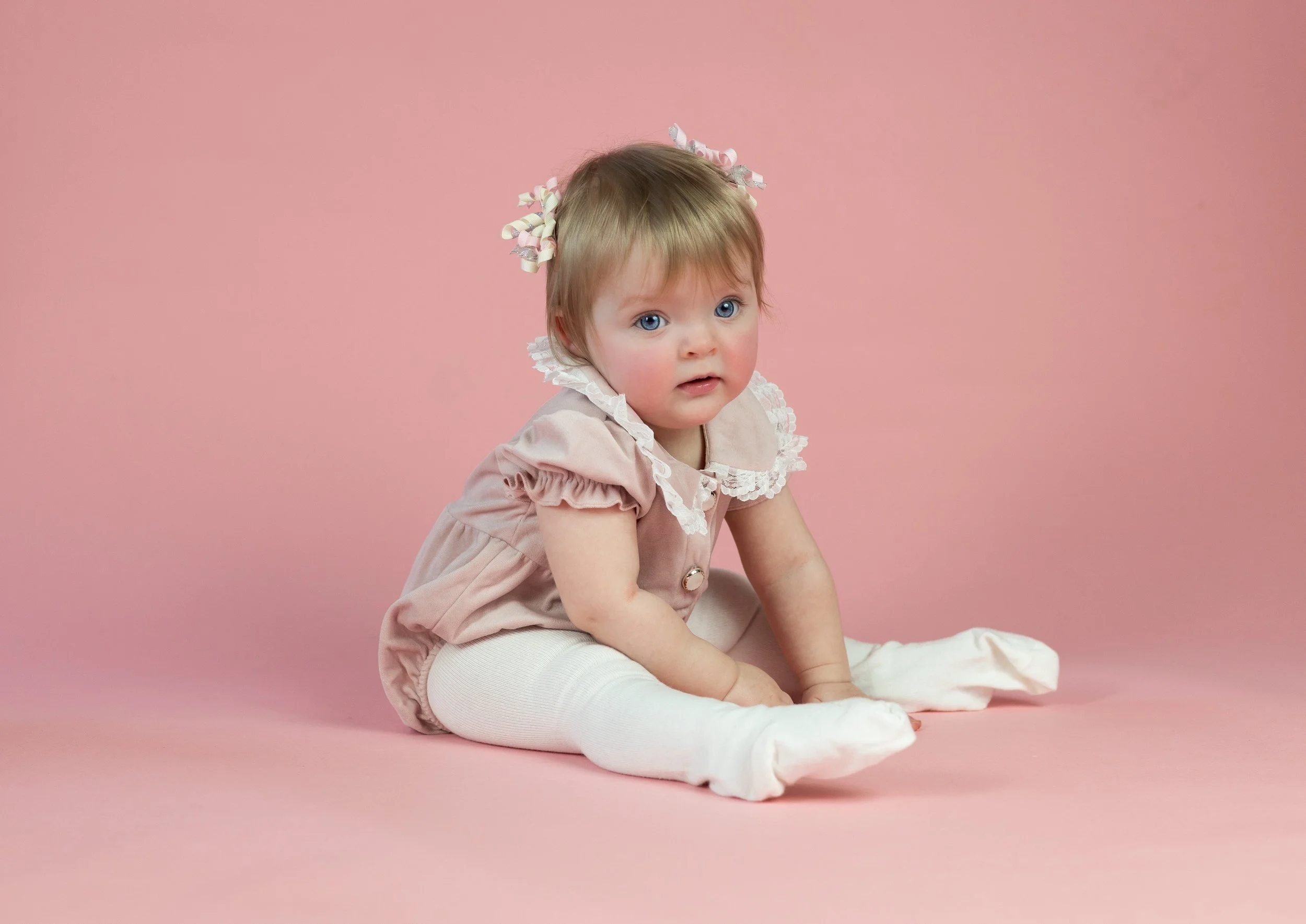 A young girl with blonde hair and blue eyes, wearing a pink dress with lace details and white tights, sitting on a pink background with a curious expression.