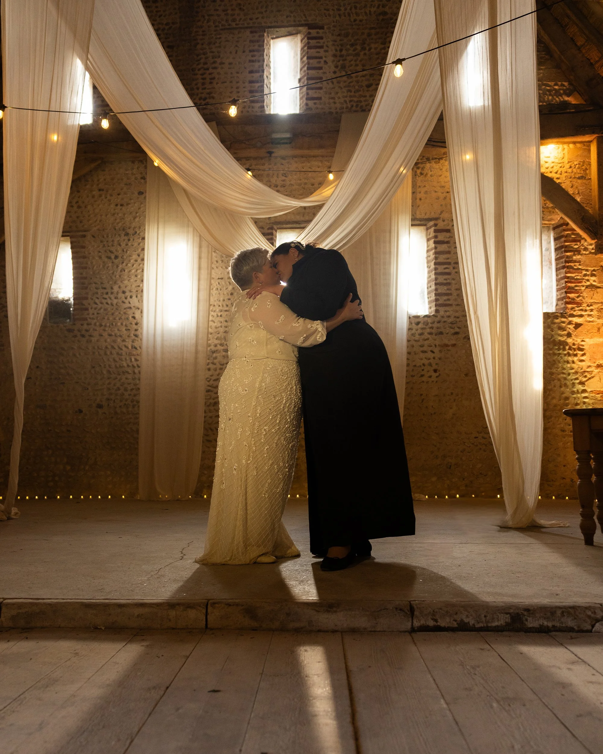 A bride and groom sharing a kiss in a rustic indoor wedding venue decorated with flowing white curtains and string lights.