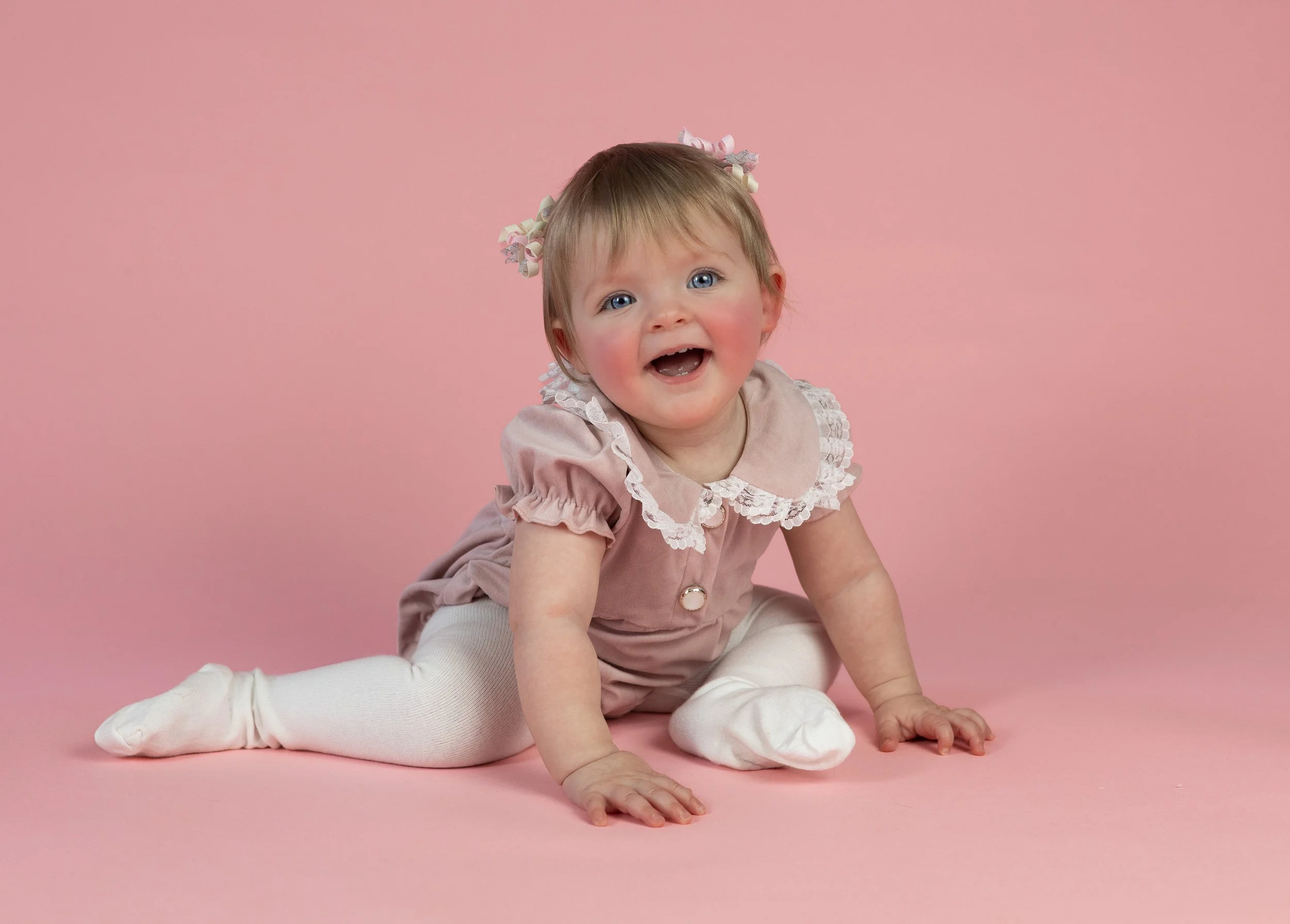 A young girl with blonde hair and blue eyes, wearing a pink dress with ruffles, white tights, and white socks, is crawling on a pink background. She has a happy expression with her mouth open and is looking at the camera.