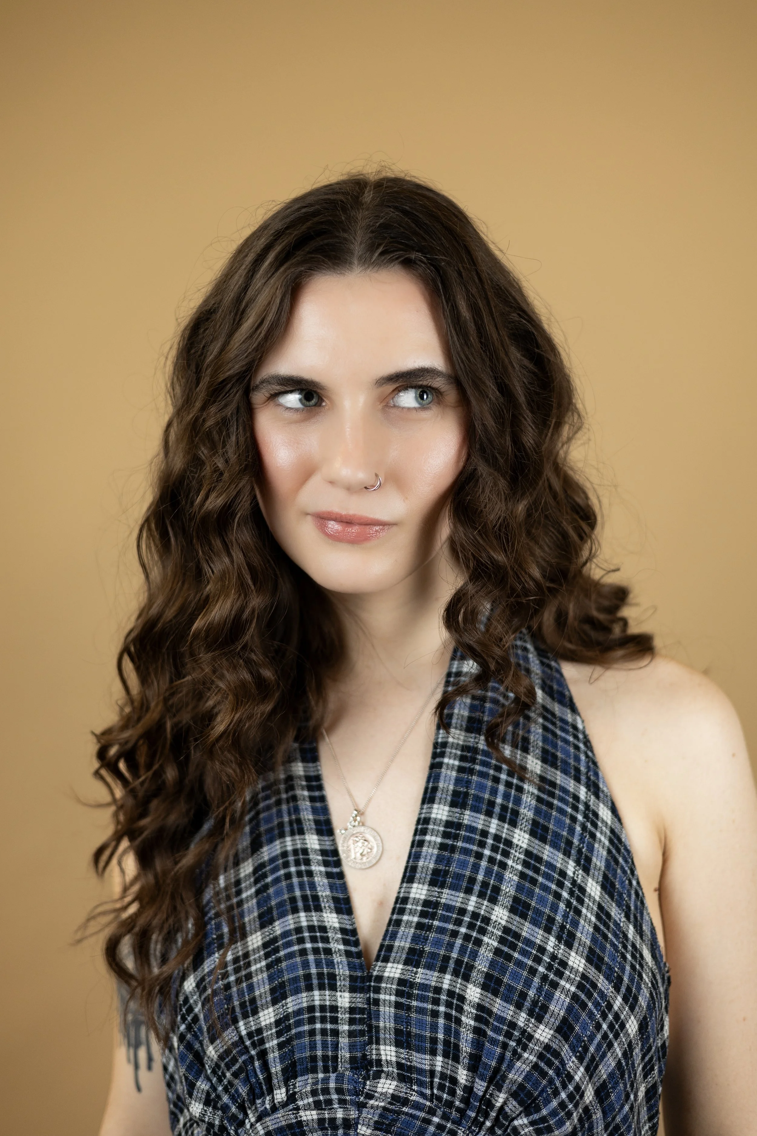 A young woman with long, curly brown hair and blue eyes looking to her right, wearing a sleeveless plaid shirt, a necklace with a round pendant, and a nose ring, against a beige background.