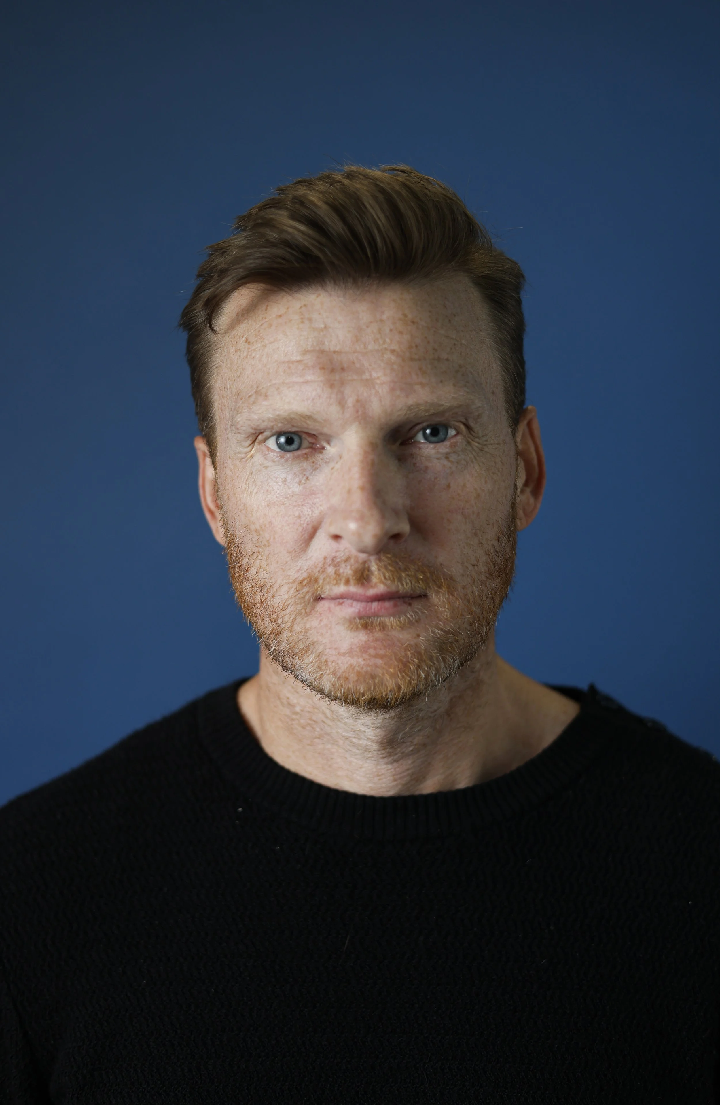 Close-up portrait of a man with short brown hair and a reddish beard wearing a black sweater against a blue background.