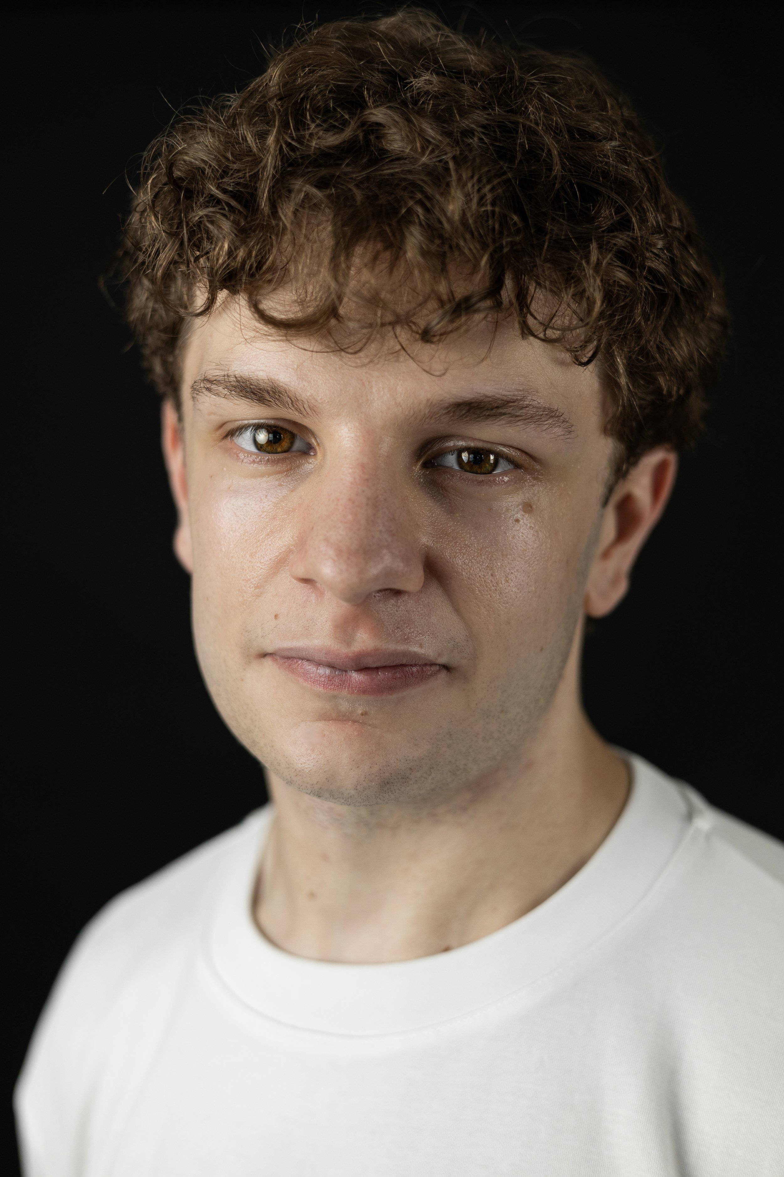A young man with curly brown hair, light skin, and brown eyes, wearing a white t-shirt, standing against a black background.