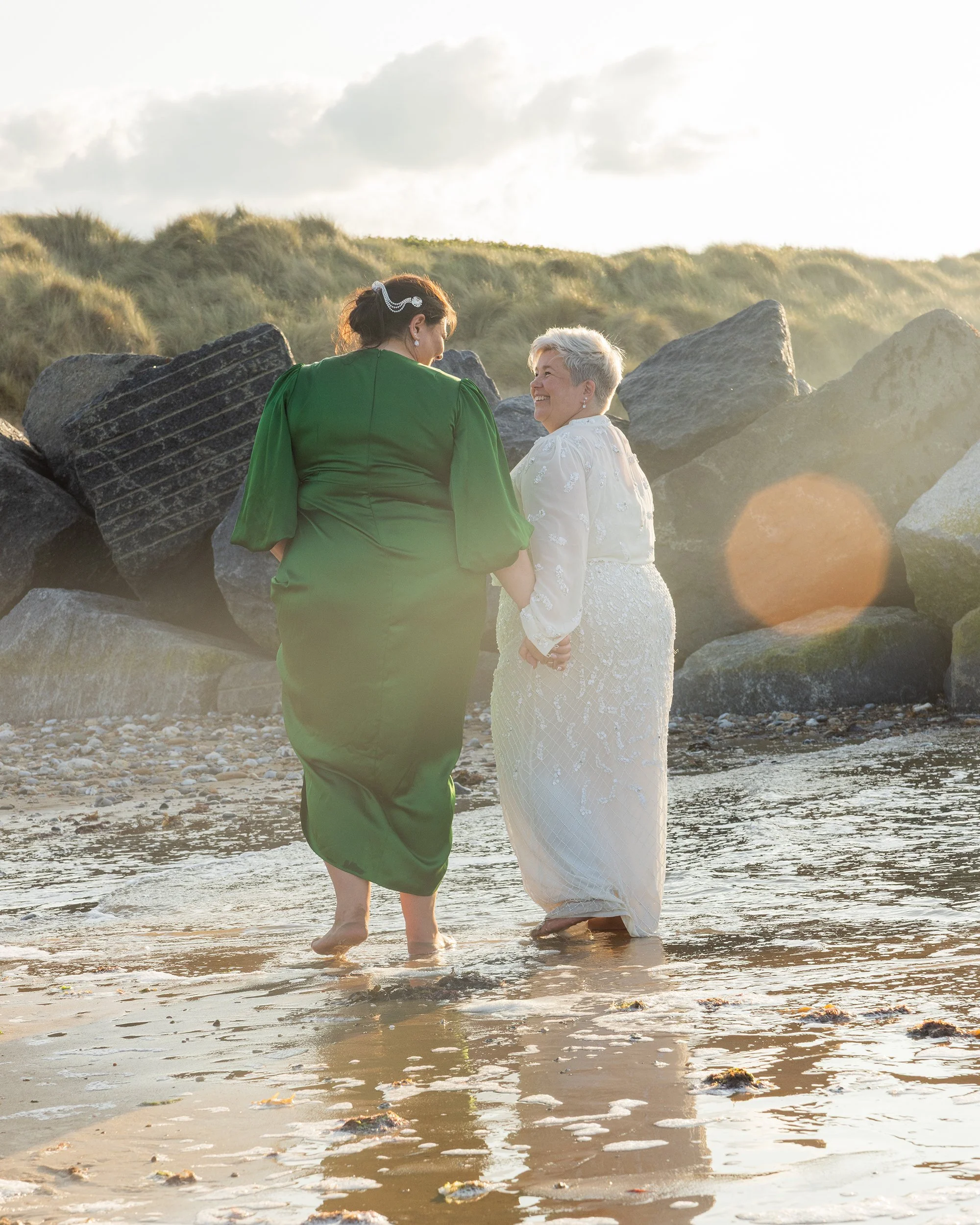 Two women holding hands walking in shallow ocean water near rocks at sunset or sunrise, with grassy dunes and cloudy sky in the background.