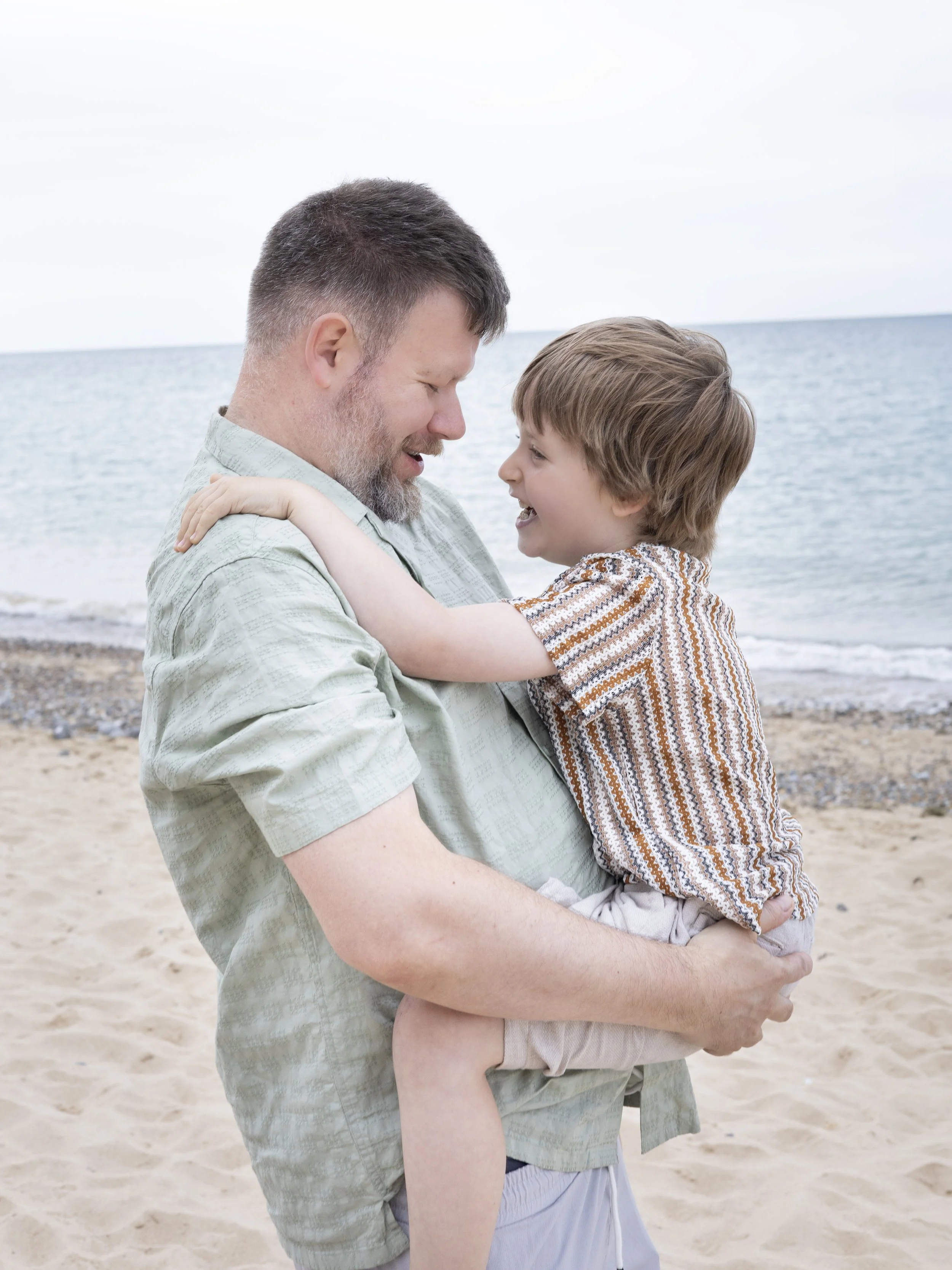 A man carrying a young boy on a beach, smiling at each other with the ocean behind them.