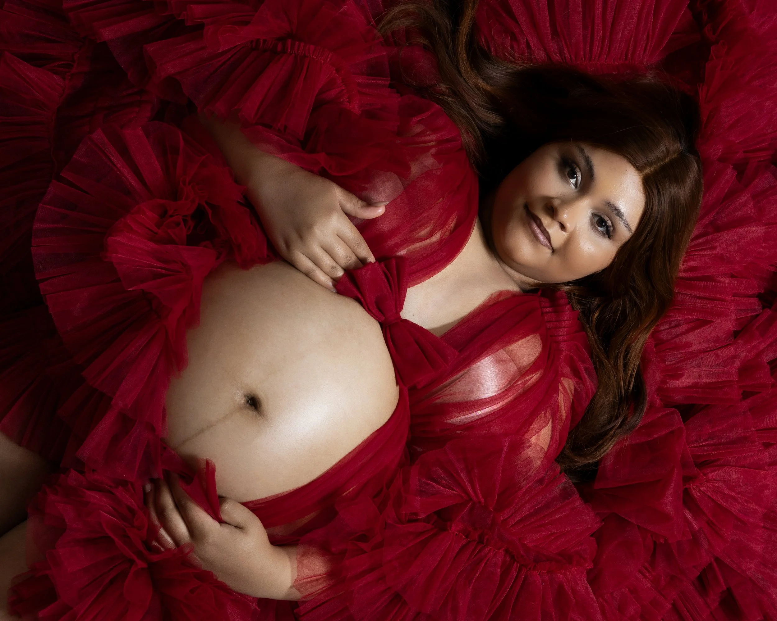 A woman with long brown hair lying on her back on a blanket of red fabric with ruffles, wearing a matching red dress, looking at the camera with a slight smile.