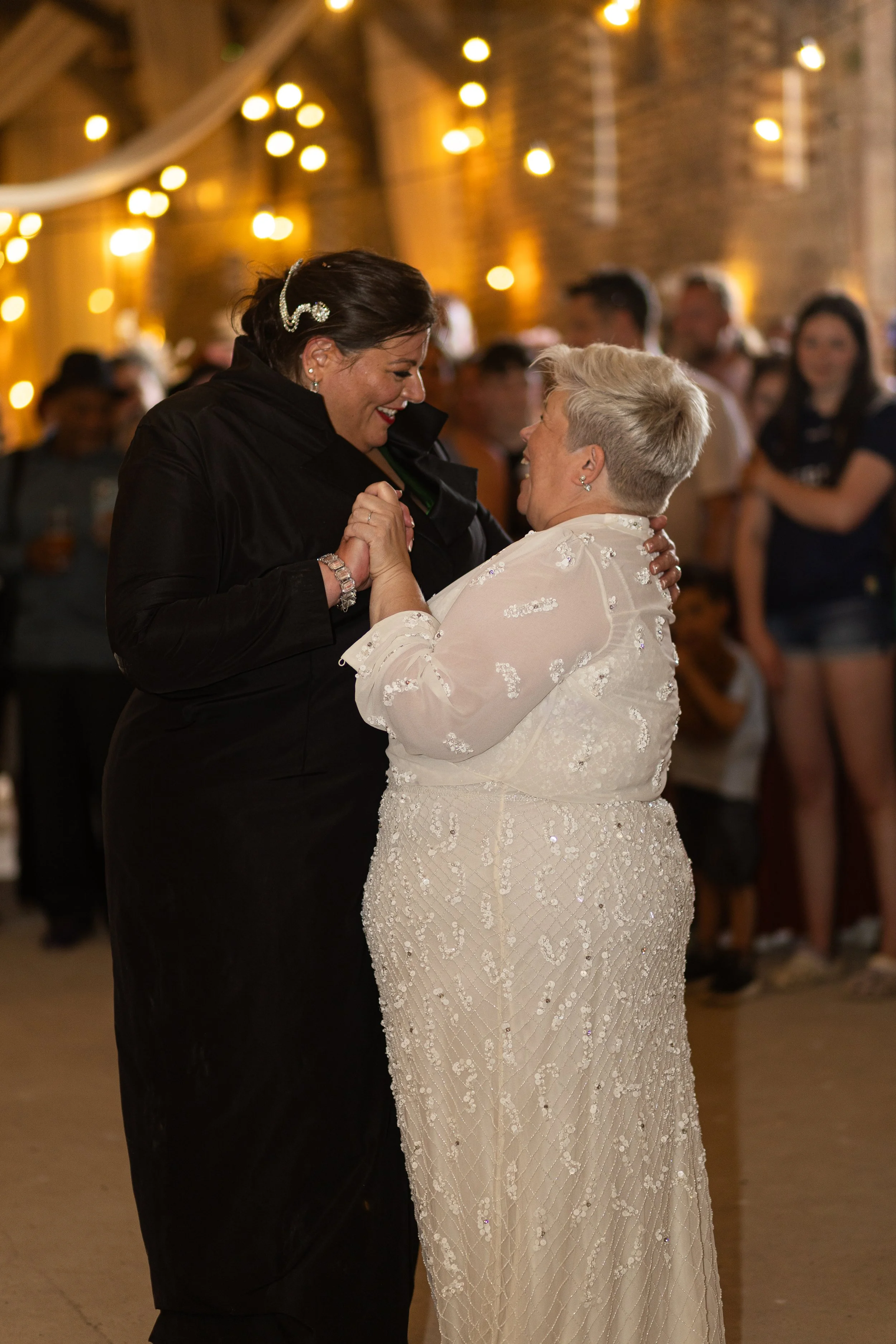 Two women dancing together at a celebration or wedding reception, smiling and holding hands, with warm string lights and a brick wall in the background.