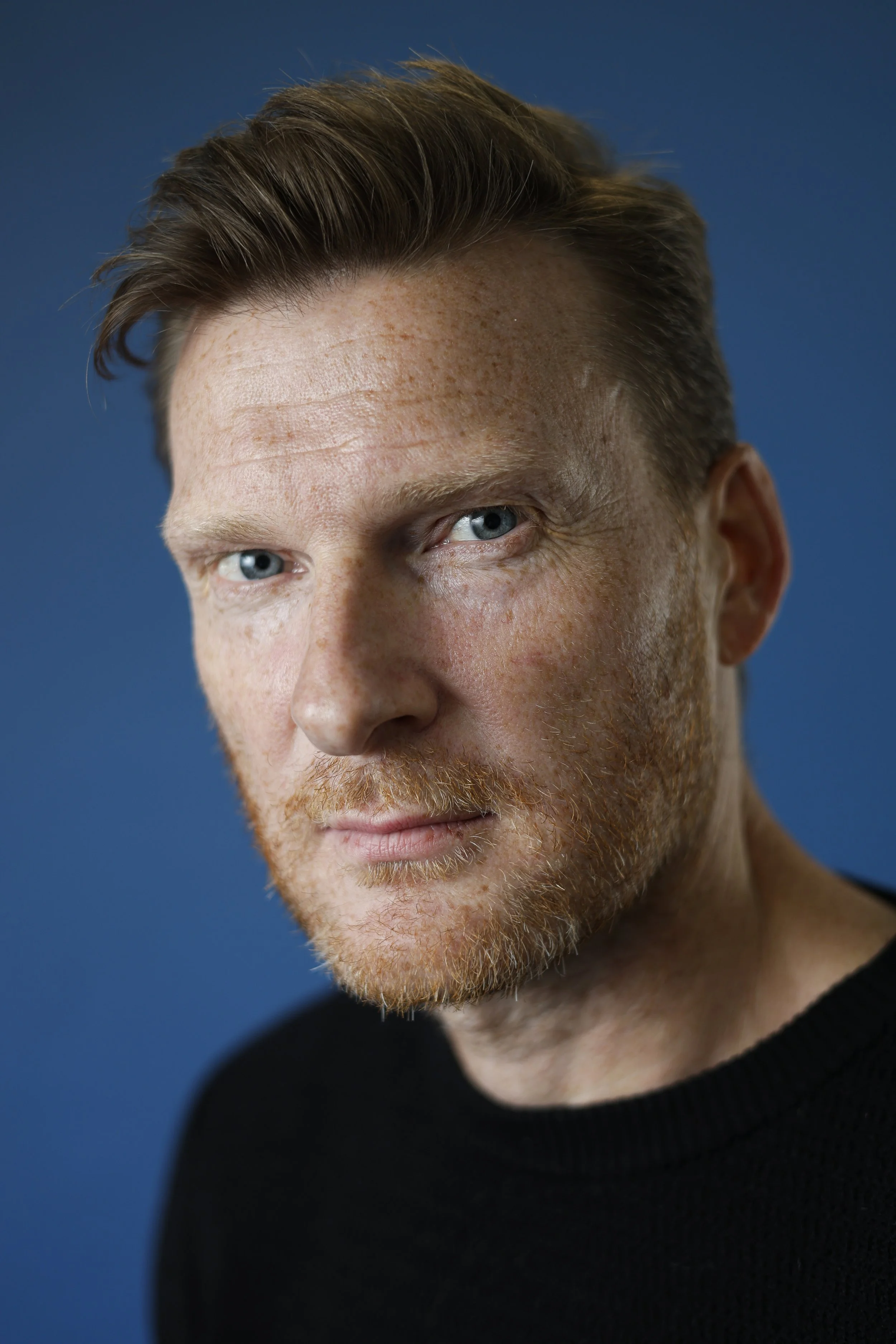Close-up portrait of a man with reddish-brown hair, blue eyes, and facial hair, wearing a black shirt, against a solid blue background.