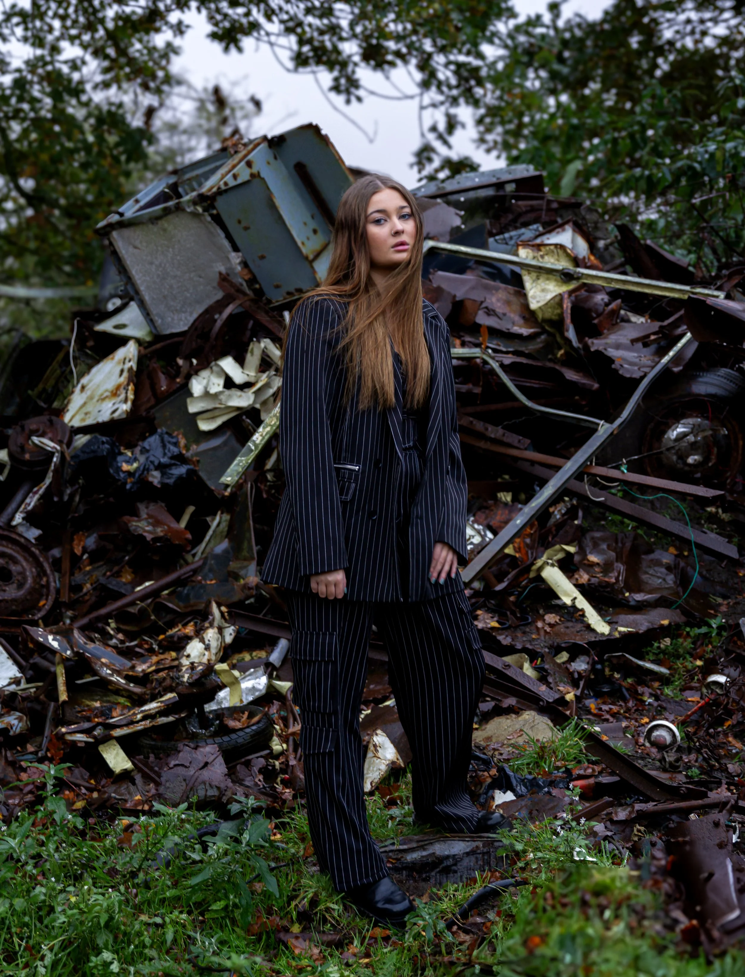 A woman with long hair wearing a black pinstripe suit stands in front of a pile of rusted scrap metal and debris outdoors.