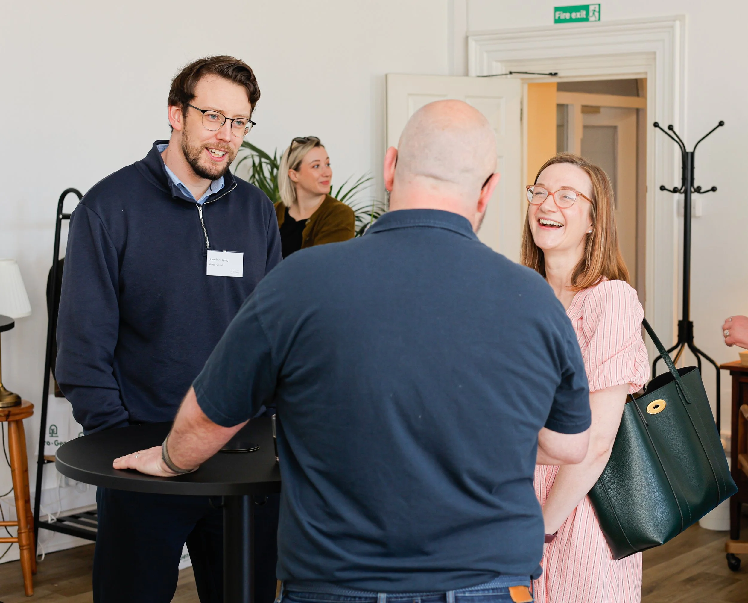 People engaging in conversation at a social or professional event, standing around a small round table in a well-lit room with a fire exit sign visible.