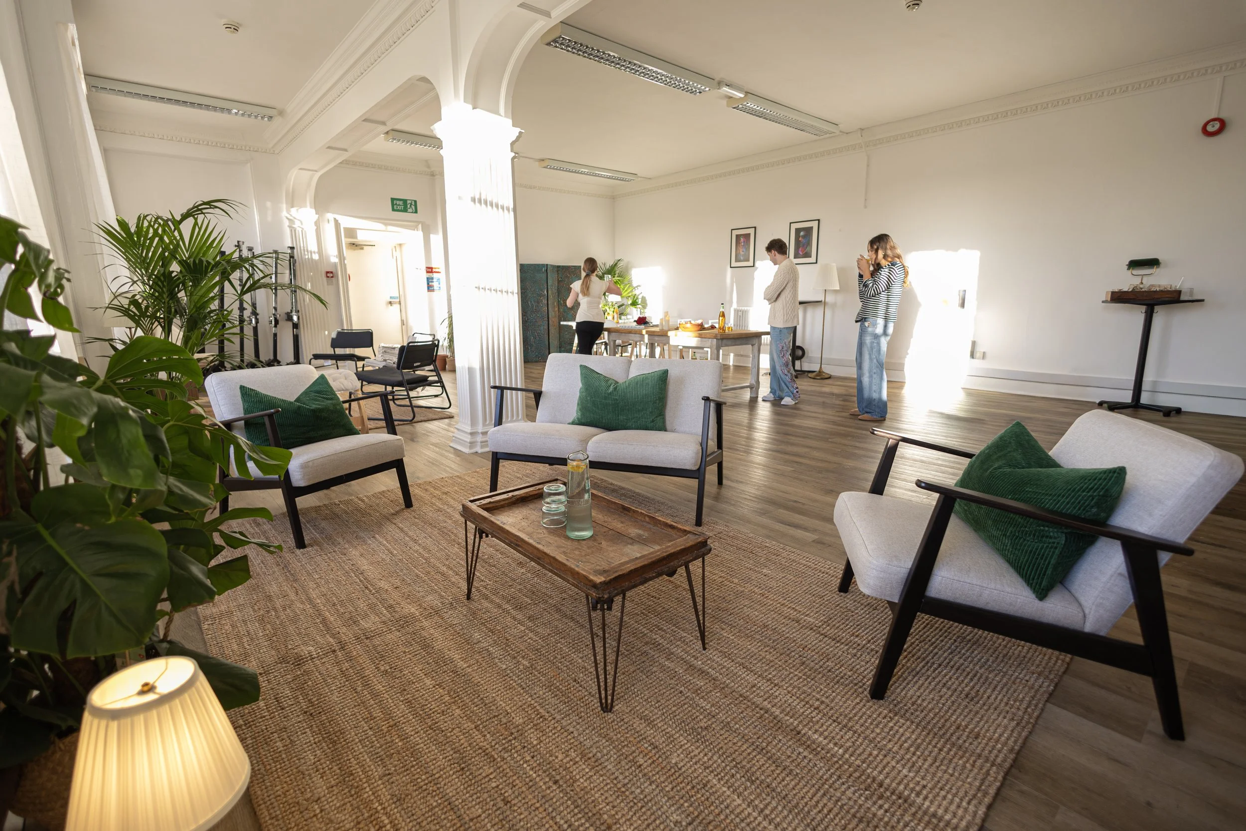 A spacious, well-lit room with white walls and wooden floors. The room is furnished with white and green armchairs, a small wooden coffee table, and some potted plants. There are three people gathering near a table with snacks and drinks, some taking