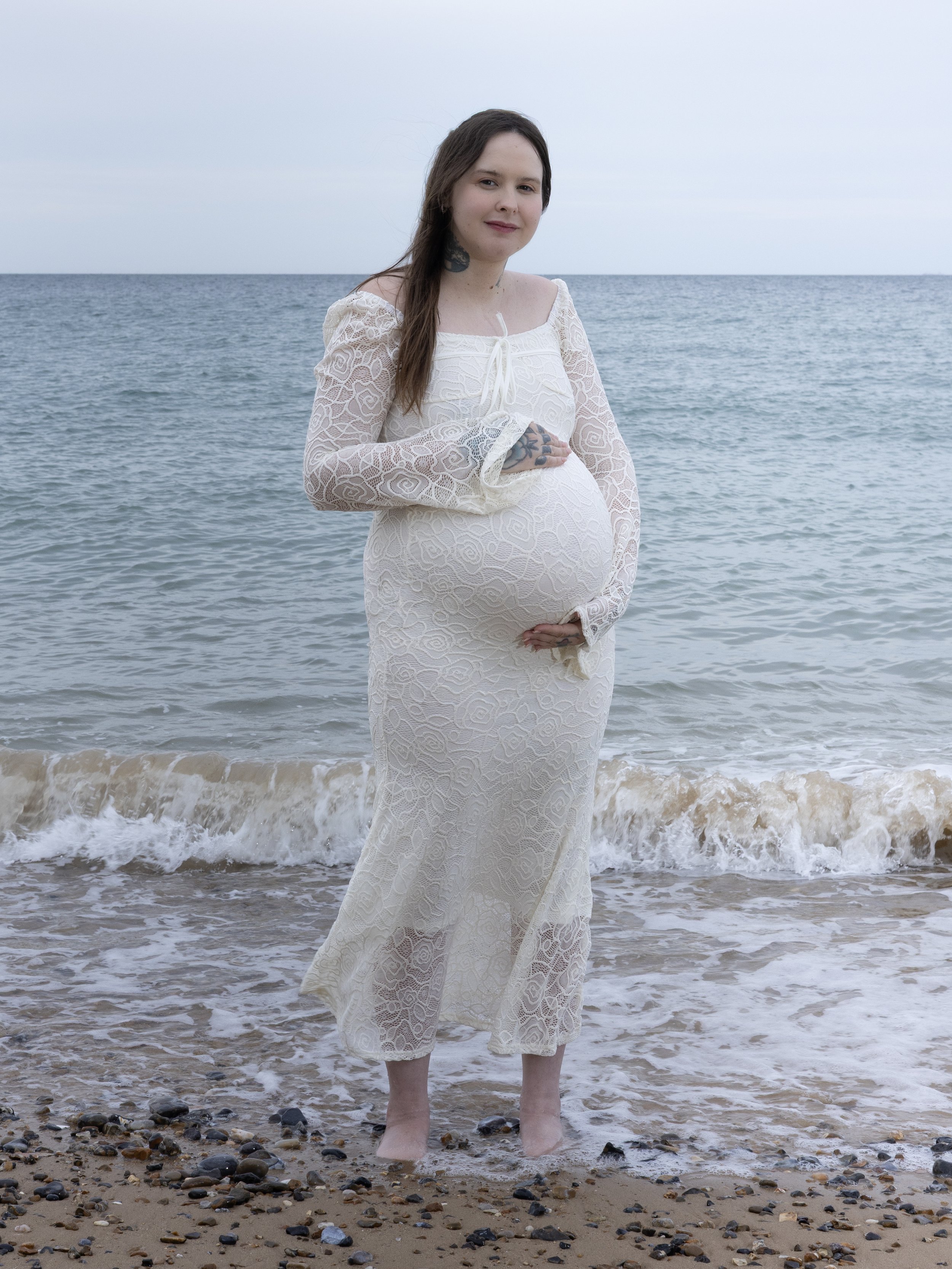 A pregnant woman in a lace white dress stands in the shallow waters of a beach with small rocks, looking at the camera with a gentle smile.