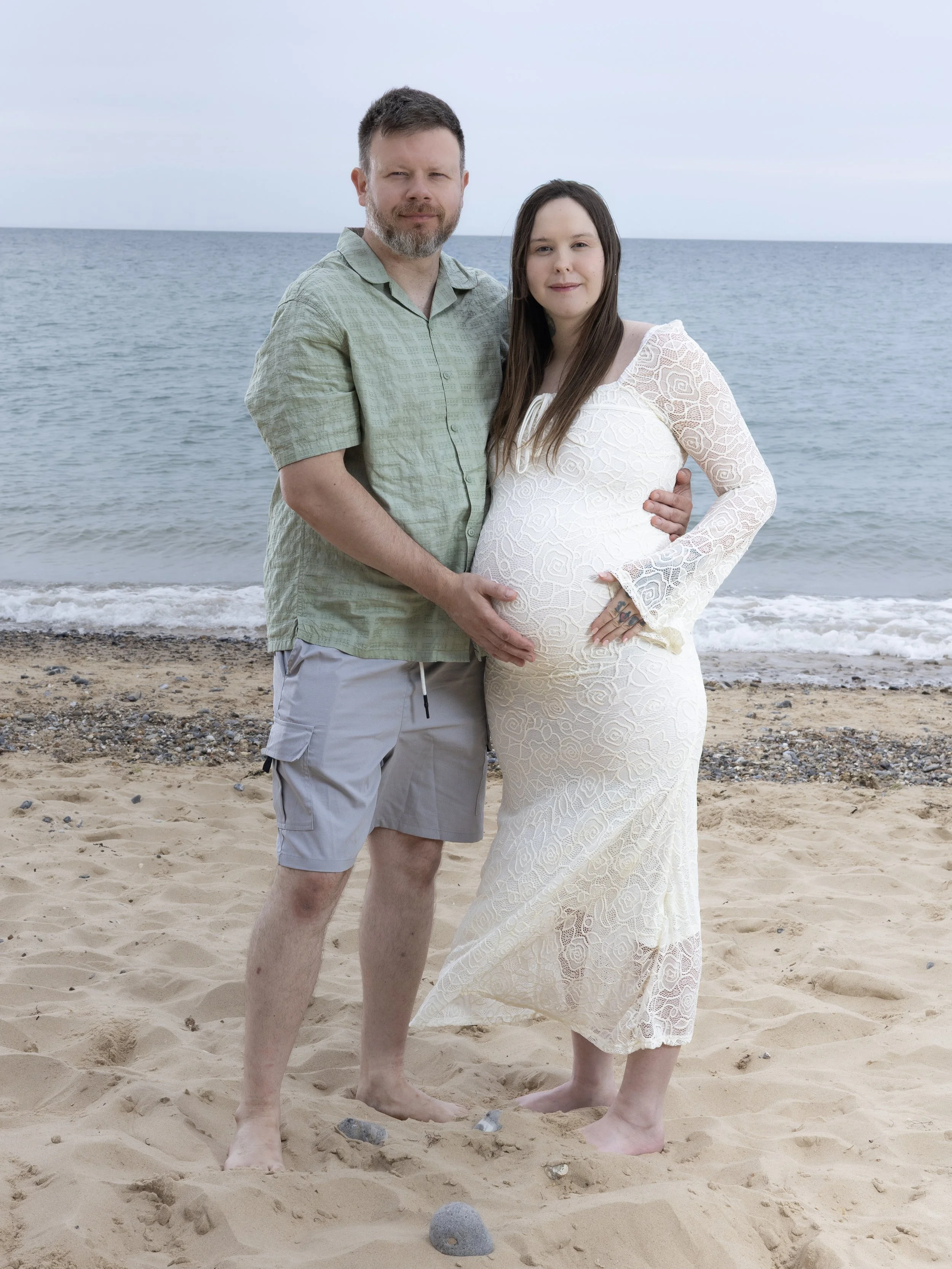 A man and a woman standing on a sandy beach next to the ocean, with the man's arm around the woman and both smiling at the camera.