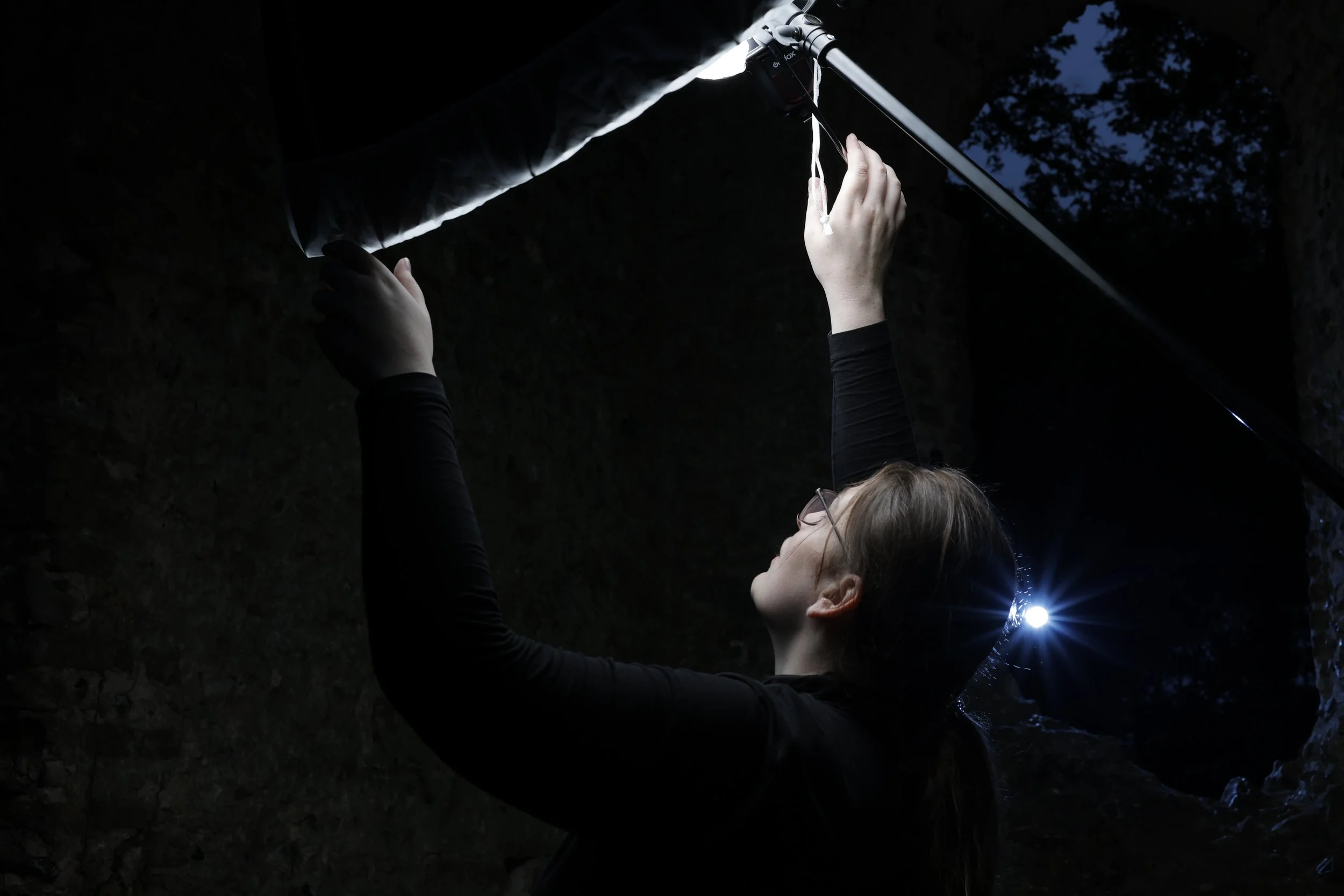 A person using a headlamp outdoors at night, reaching upward to adjust or inspect something overhead.