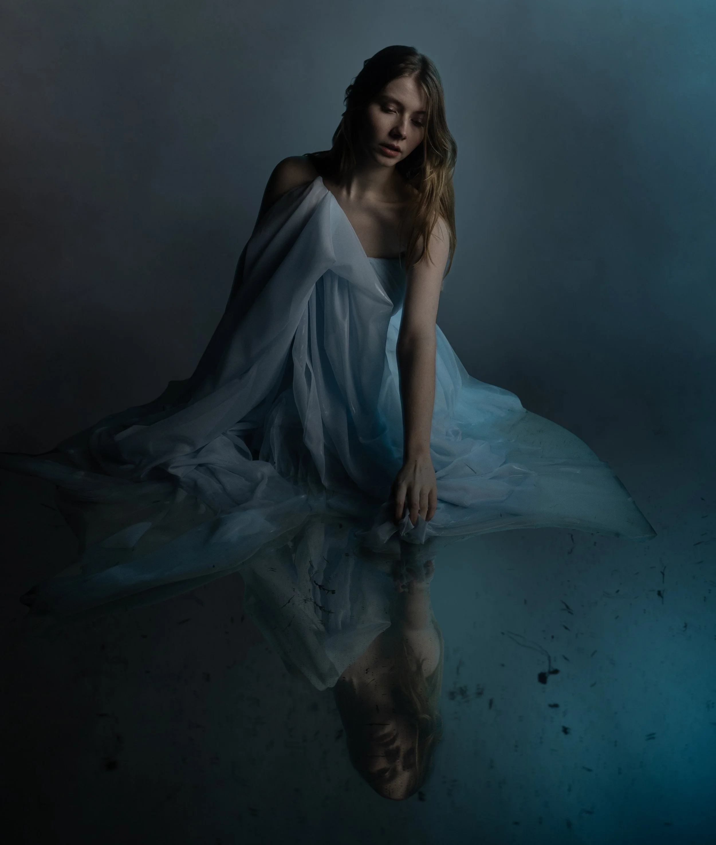 A woman with long hair, dressed in a flowing white gown, sitting on a dark ground with a somber expression, against a moody, dark background, with her reflection visible on the surface beneath her.
