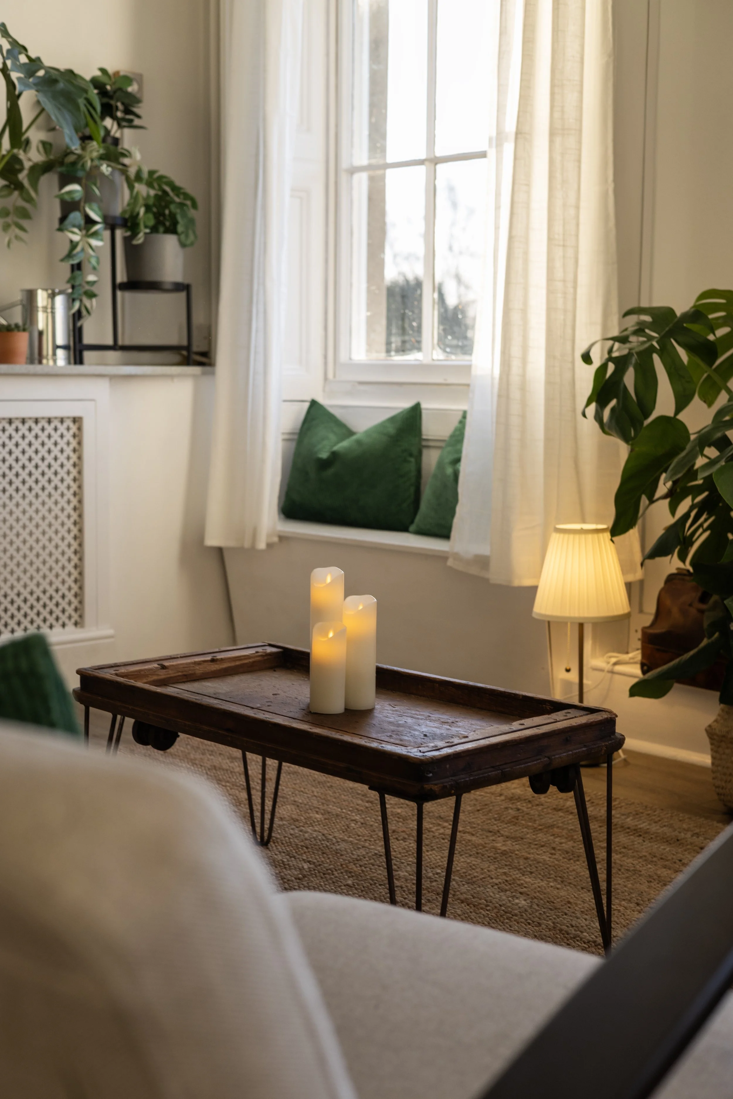 Cozy living room with a window seat with green pillows, a wooden coffee table with candles, a beige lamp, and lush green indoor plants near the window with cream curtains.