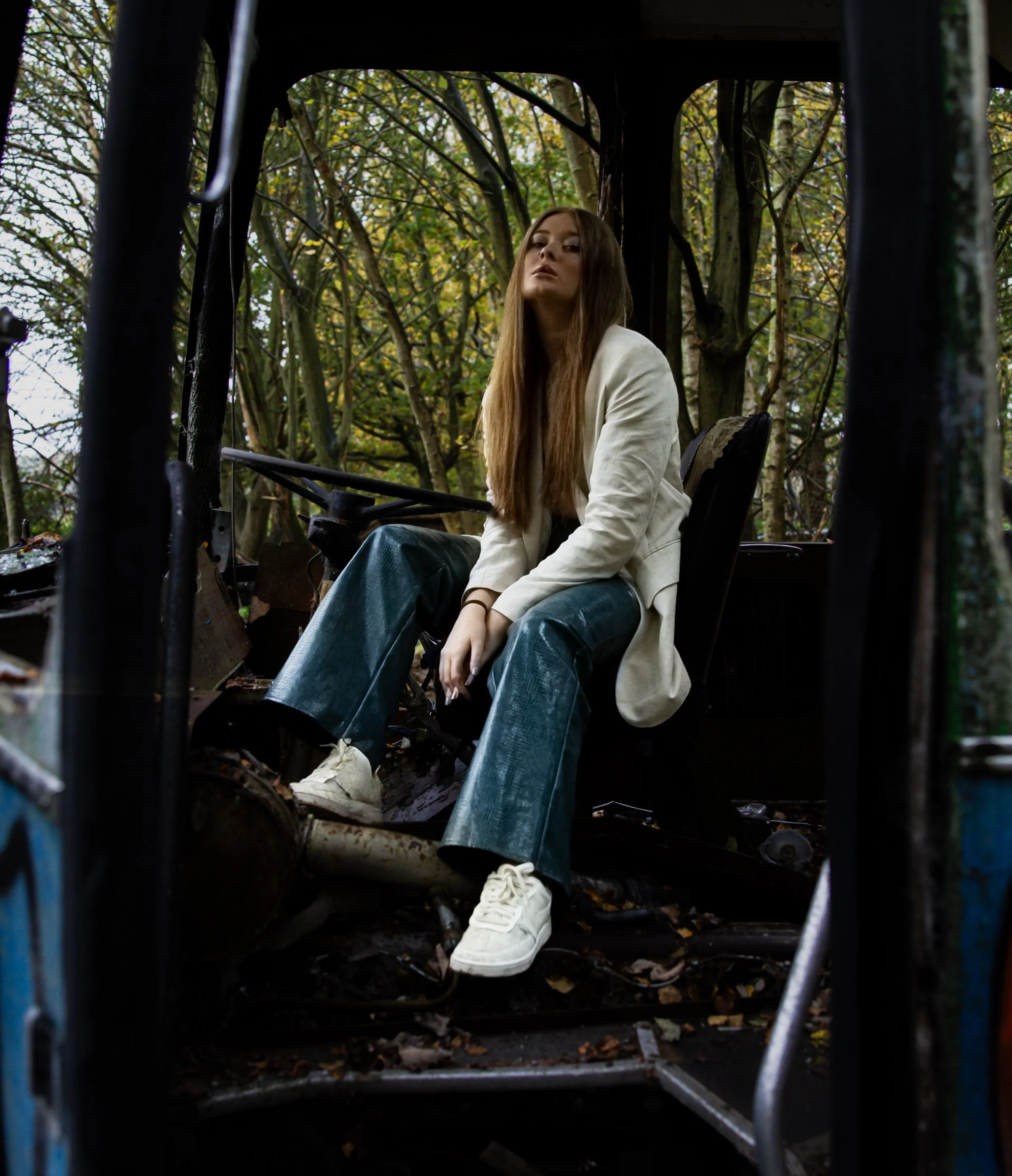 A young woman with long brown hair wearing a white jacket and blue pants sitting in the back of a trash truck surrounded by autumn leaves and trees.