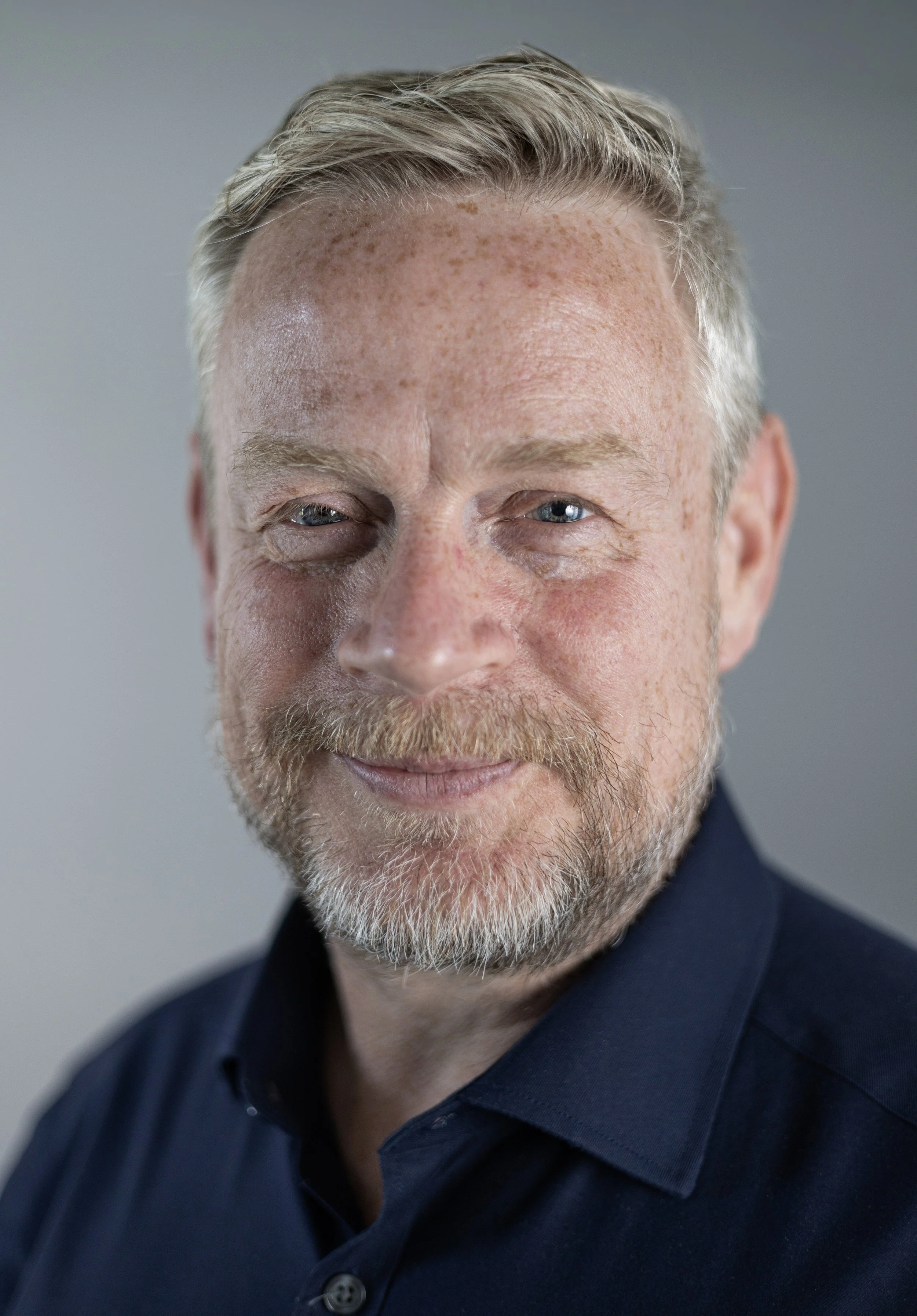 Close-up portrait of a middle-aged man with gray hair, blue eyes, and a beard, wearing a dark blue shirt, smiling softly against a neutral background.