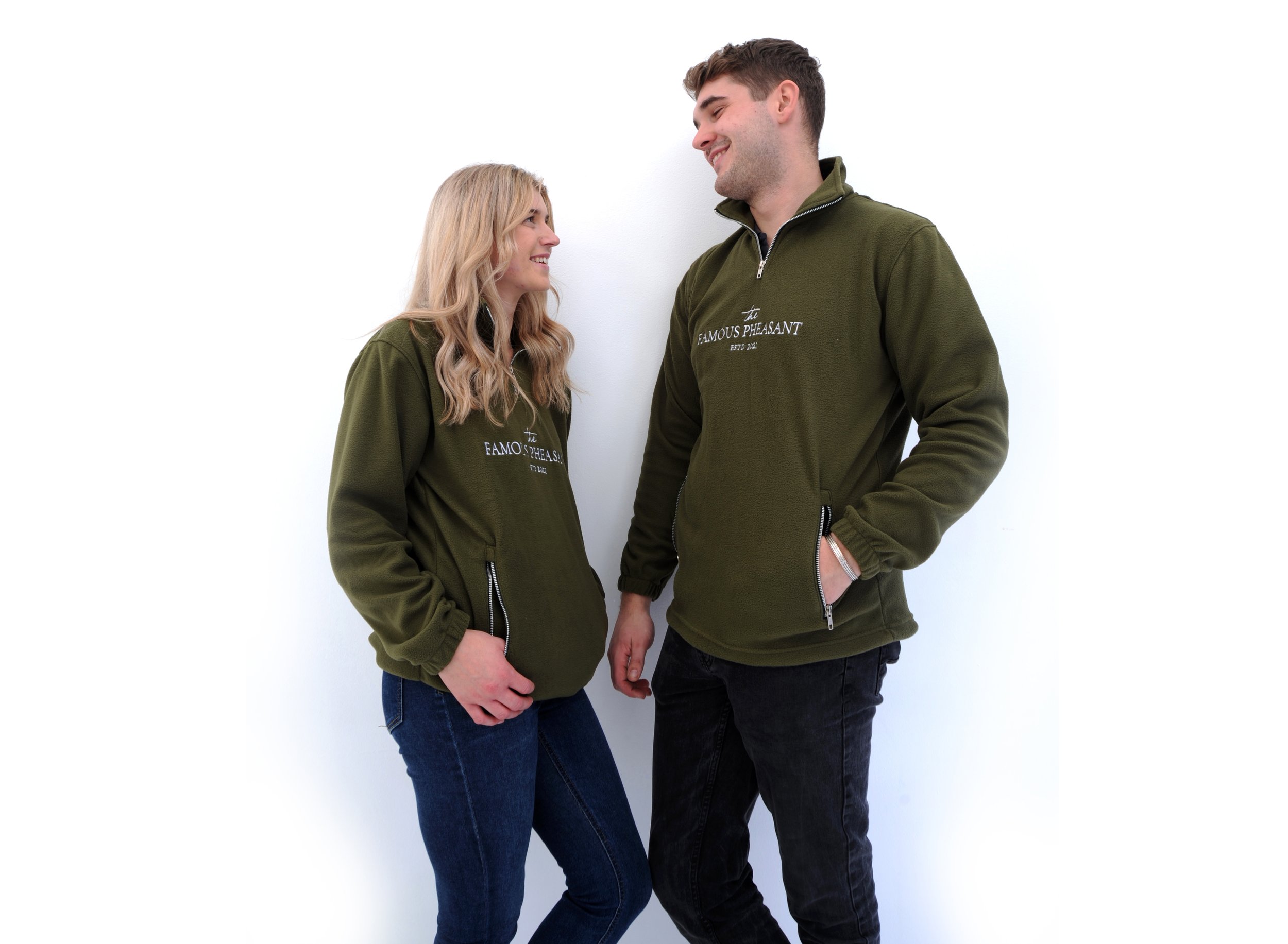 A smiling young man and woman stand together against a white background, both wearing matching green zippered sweatshirts with the phrase "the FAMOUS PHEASANT" embroidered on them.