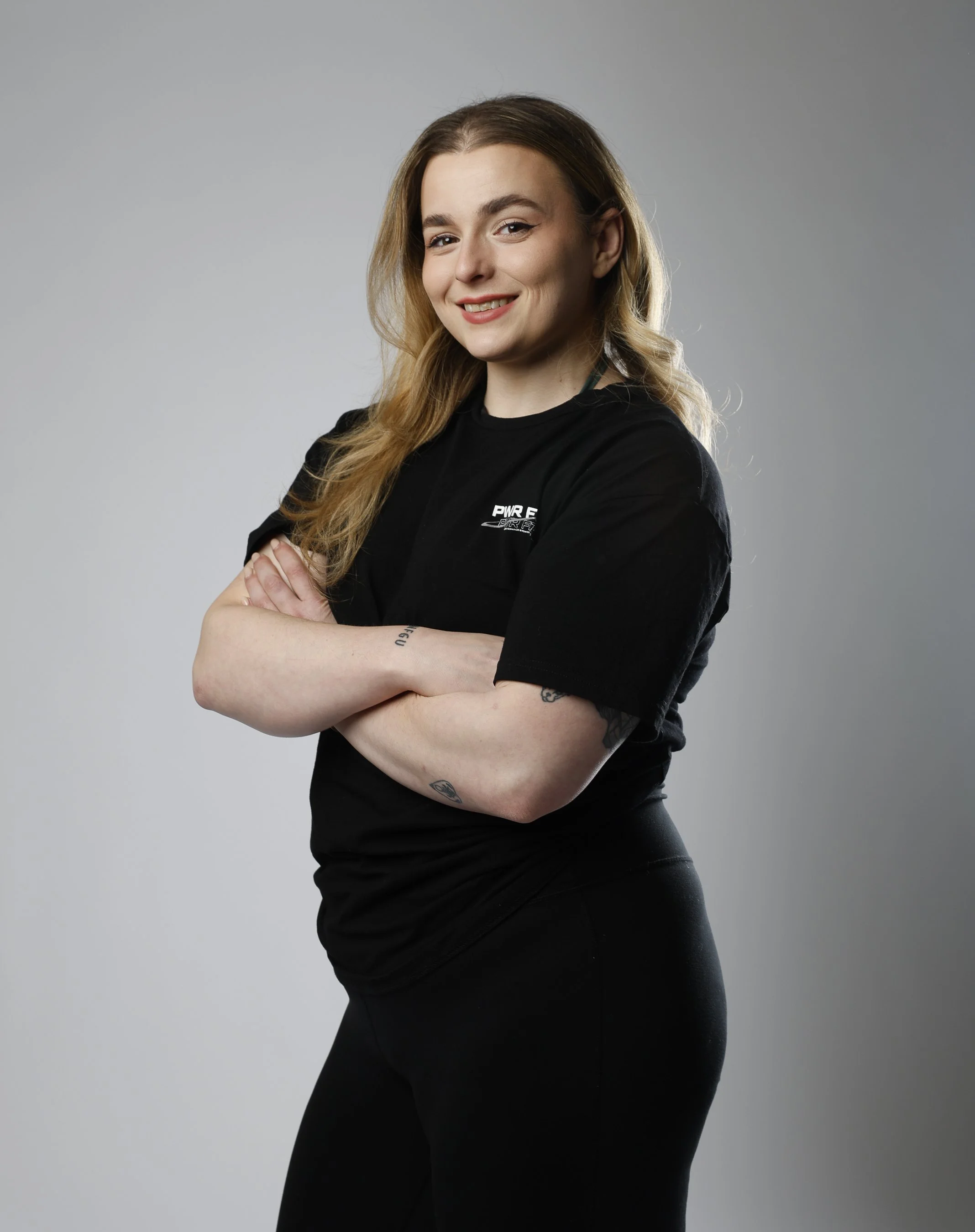 A woman with long light brown hair, smiling, wearing a black t-shirt and black leggings, standing with arms crossed against a plain gray background.