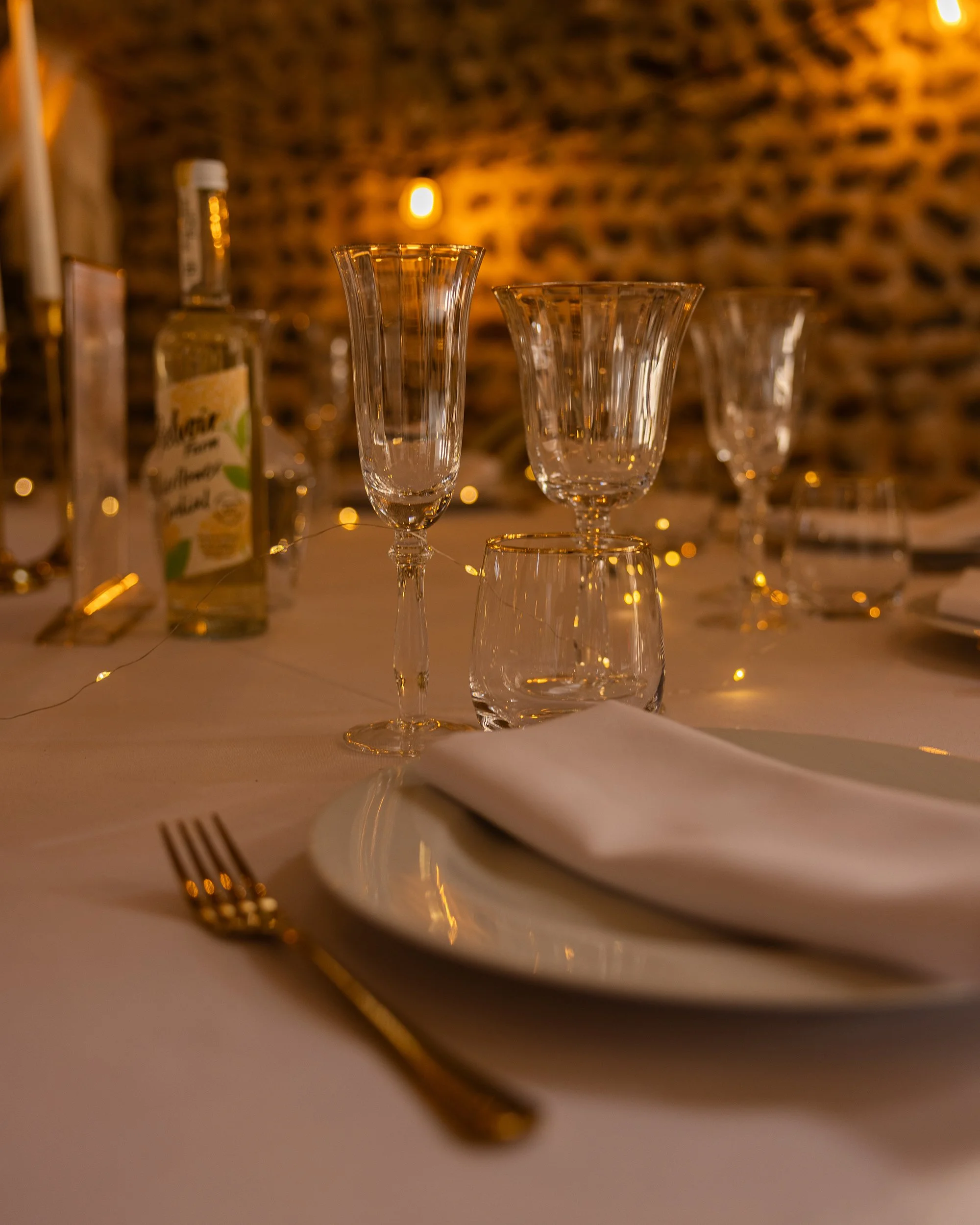 Upscale photo of a set dining table with elegant glassware, a white plate, a folded napkin, and a gold fork on a white tablecloth, illuminated with warm lighting.