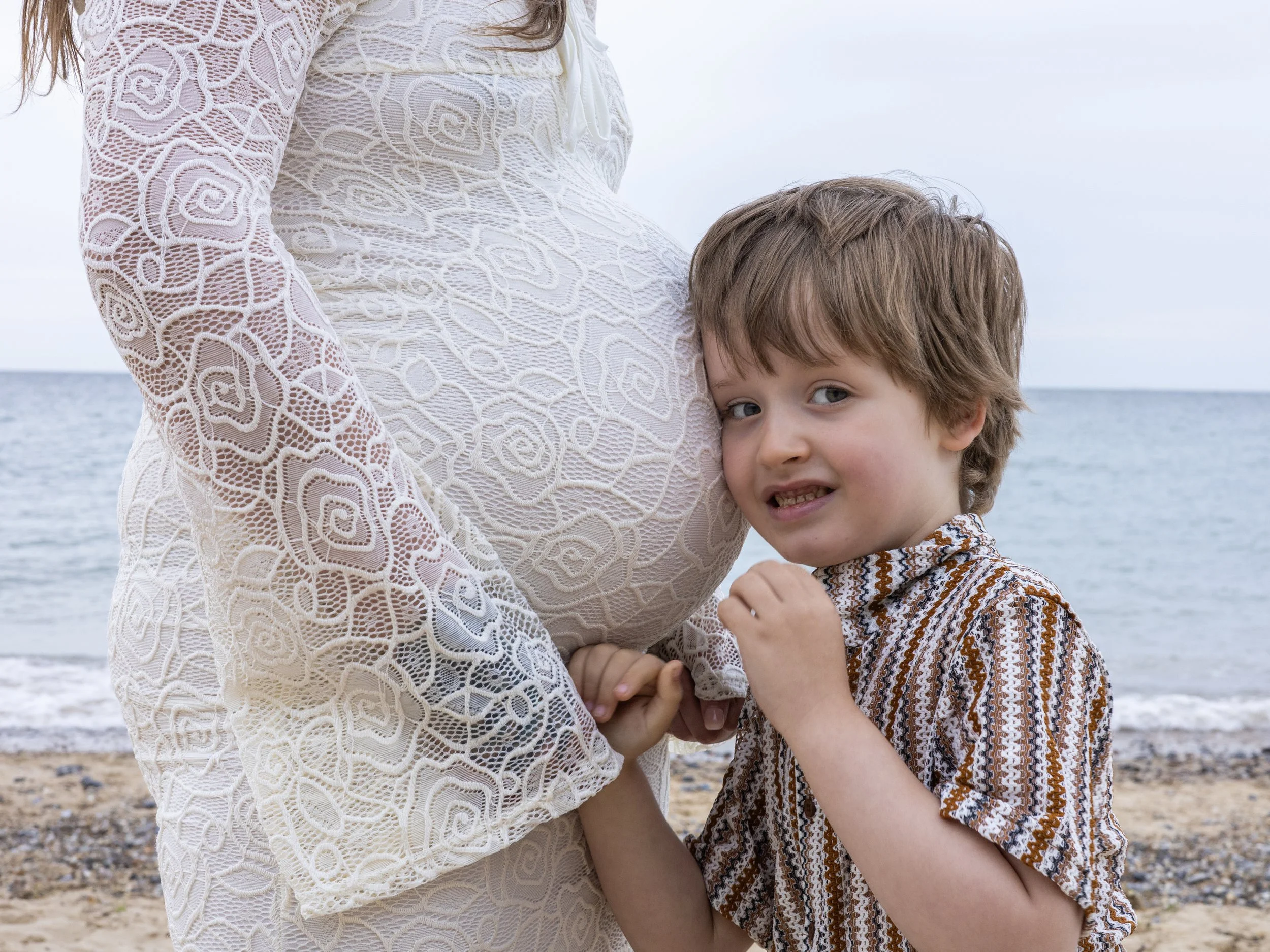 A boy with light brown hair and a patterned shirt is hugging a pregnant woman with long hair and a lace dress near the ocean.