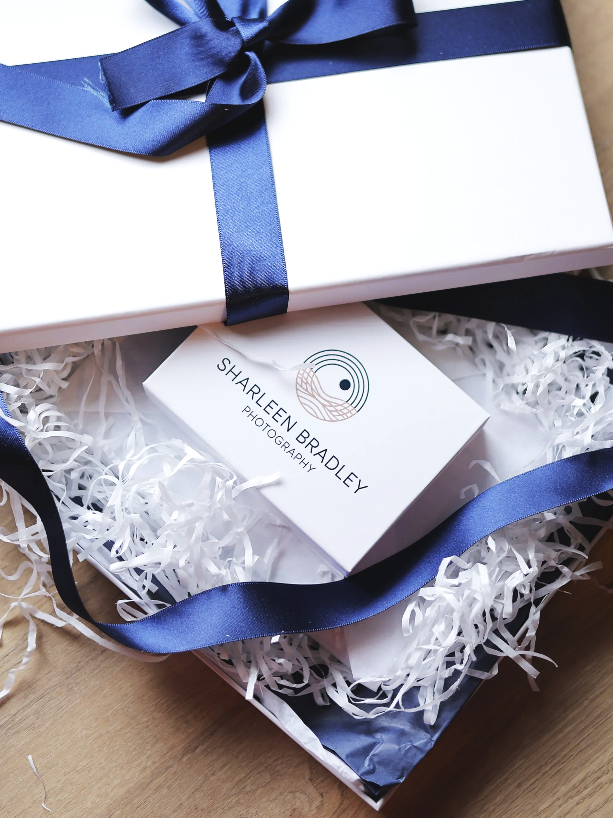 A white gift box wrapped with a dark blue satin ribbon tied in a bow, placed on white shredded paper packaging fill near a business card for 'Sharleen Bradley Photography' with a circular logo.