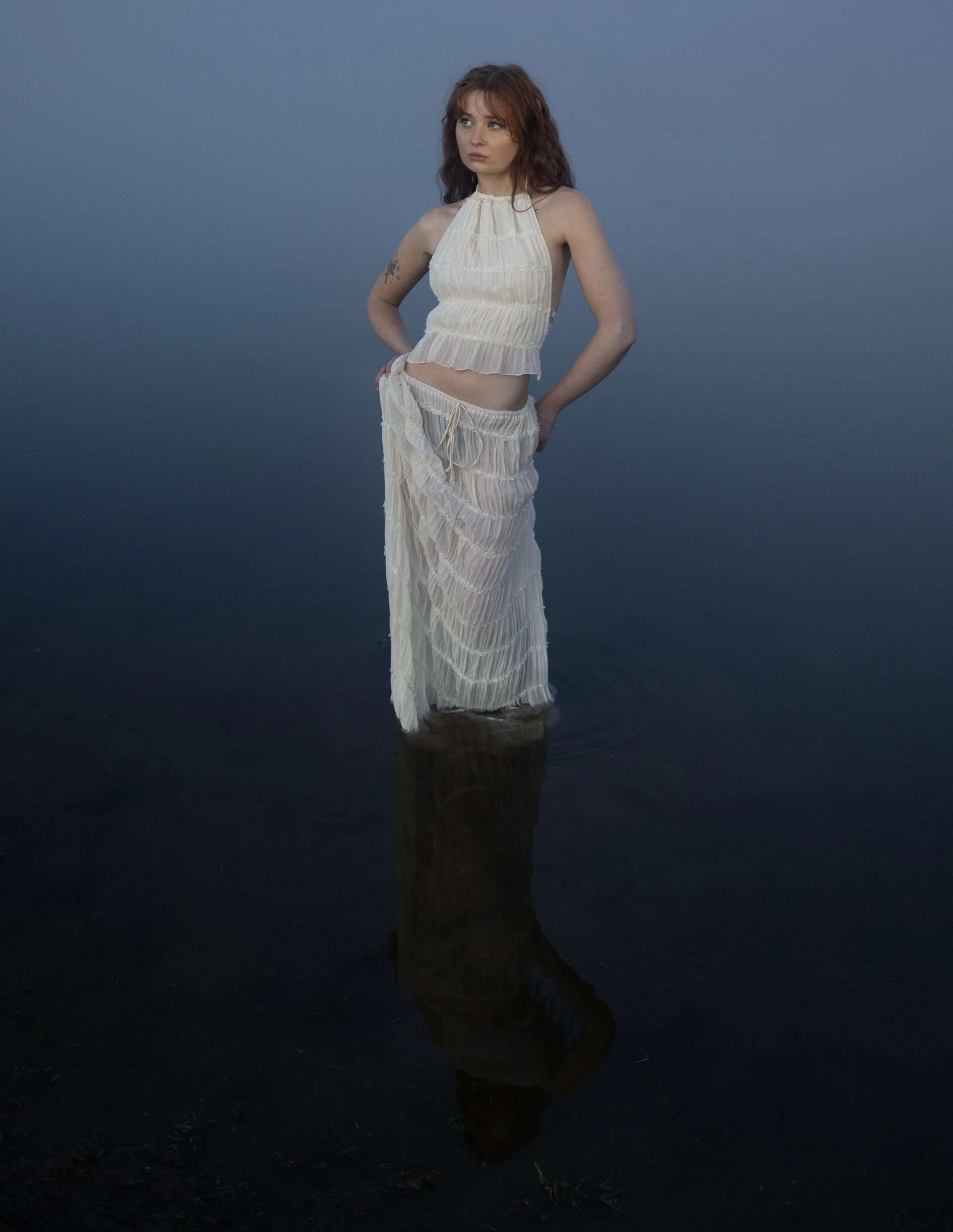A woman standing in shallow water at dusk or dawn, wearing a white, flowy, sleeveless dress, with her hands on her hips, looking to her left, with a calm and contemplative expression.