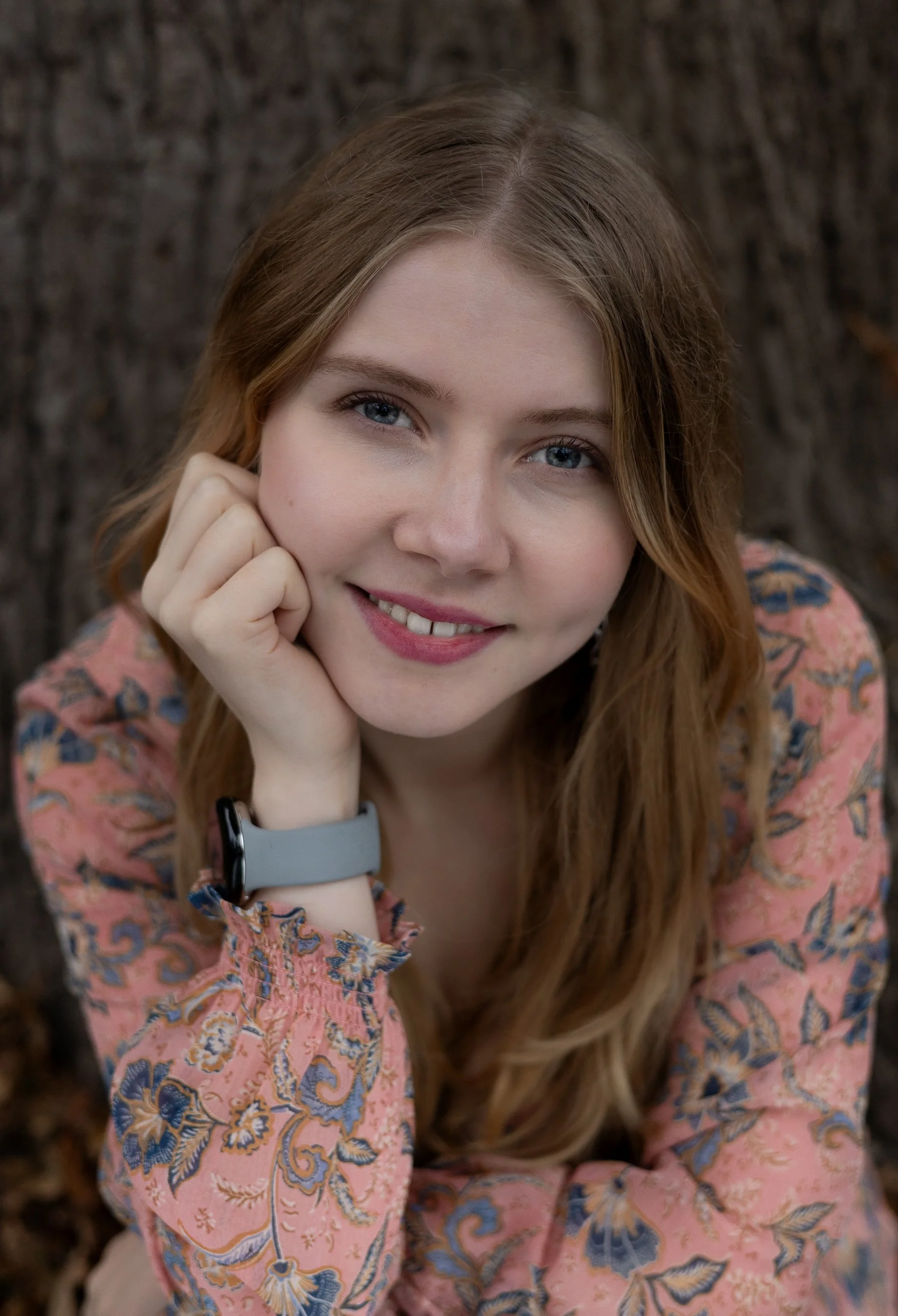 A smiling woman with blue eyes and long light brown hair resting her face on her hand, wearing a pink and blue paisley patterned shirt and a gray watch, sitting outdoors with a blurred, dark, and textured background.