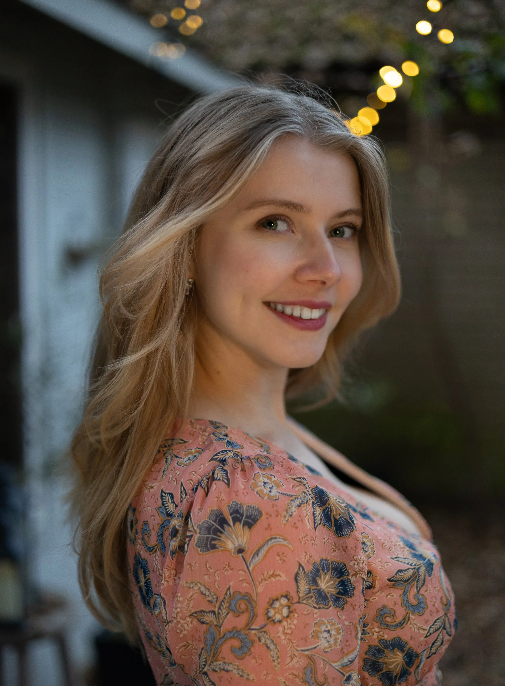 Young woman with long blonde hair, smiling, wearing a pink floral dress, outdoors during evening with string lights in the background.
