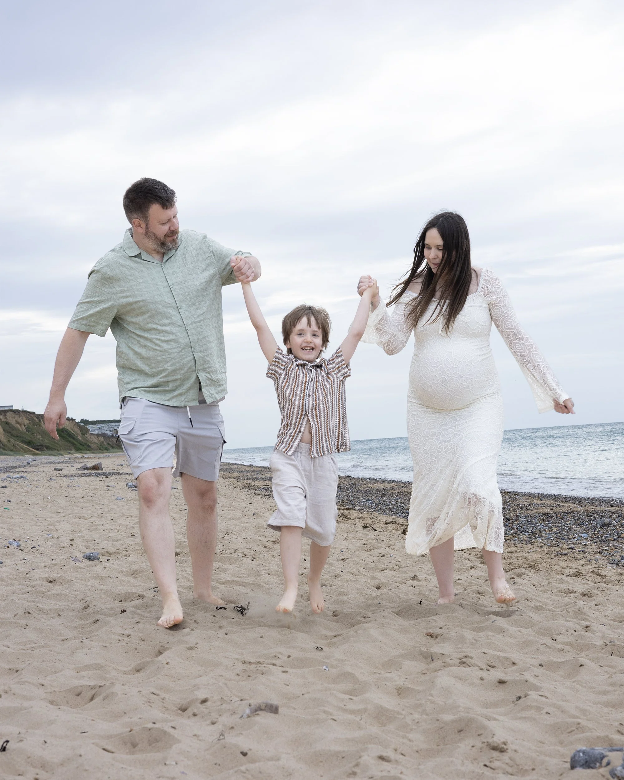 A family of three walking barefoot on a sandy beach, holding hands. The father is on the left, wearing a light green shirt and shorts. The mother is on the right, wearing a long white dress. The child, a boy, is in the middle, smiling, wearing a striped shirt and light-colored pants.