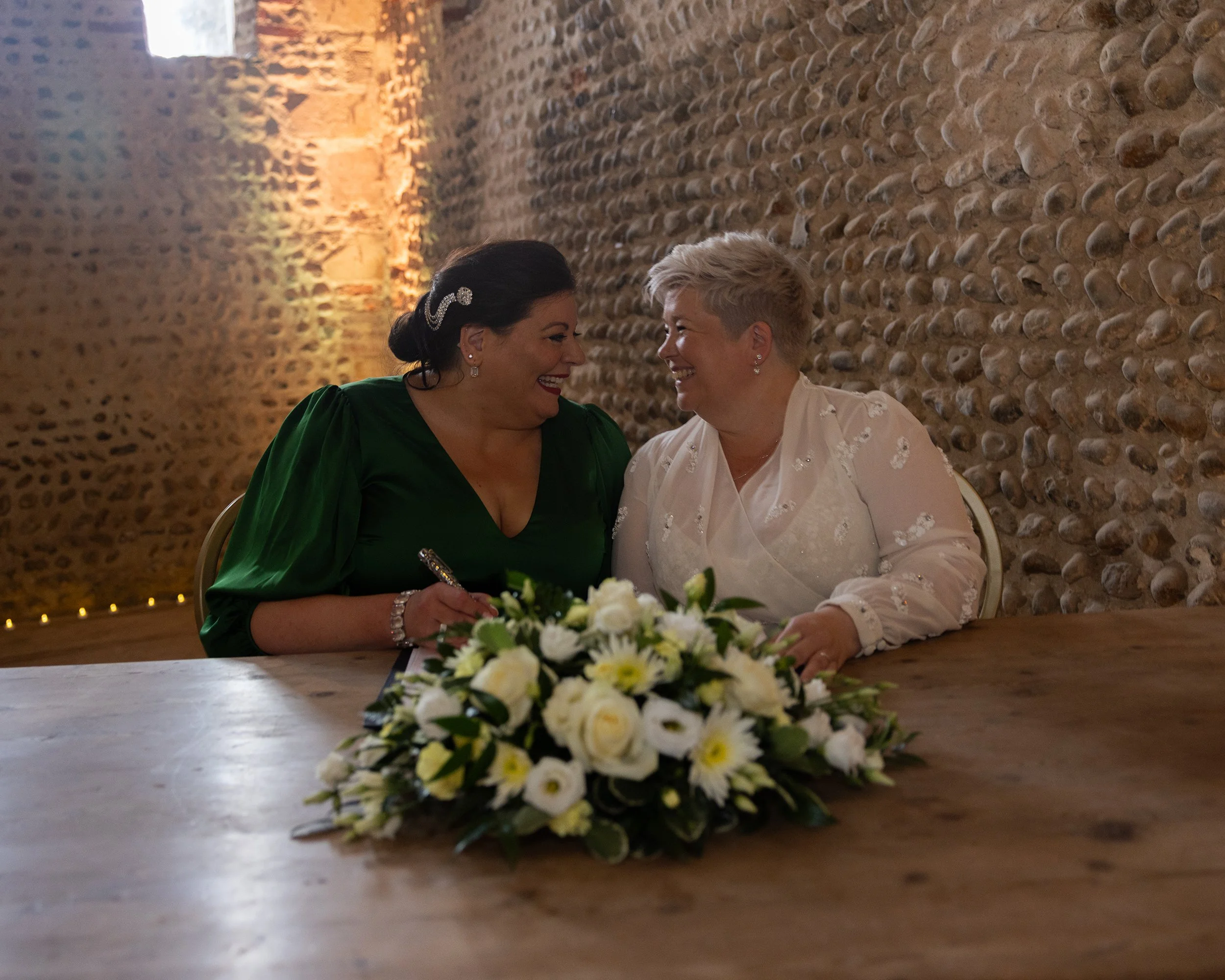 Two women, one with dark hair and the other with short blonde hair, smiling and sitting at a table with a floral centerpiece, in a room with textured brick walls and warm lighting.