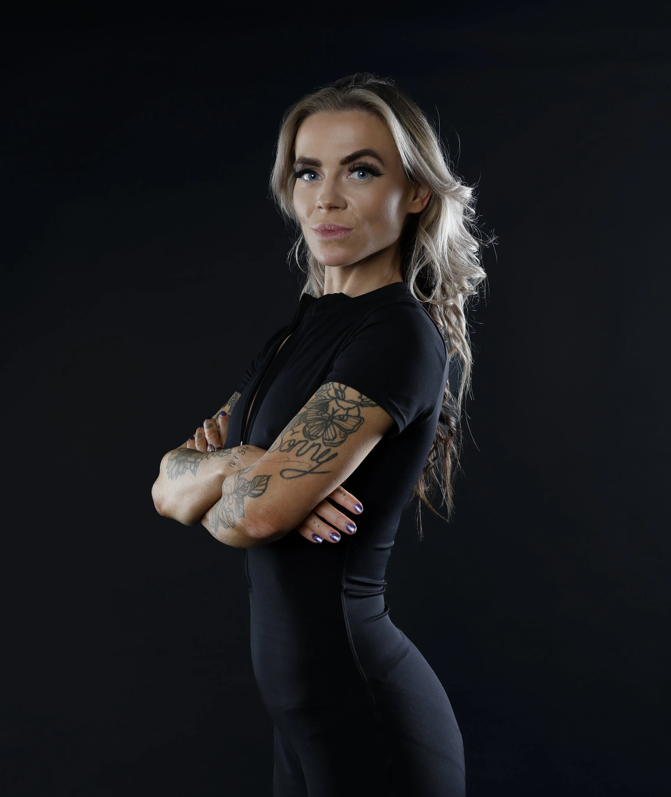 A woman with blonde hair, blue eyes, and tattoos on her arms, wearing a black top and black leggings, standing against a dark background with her arms crossed.