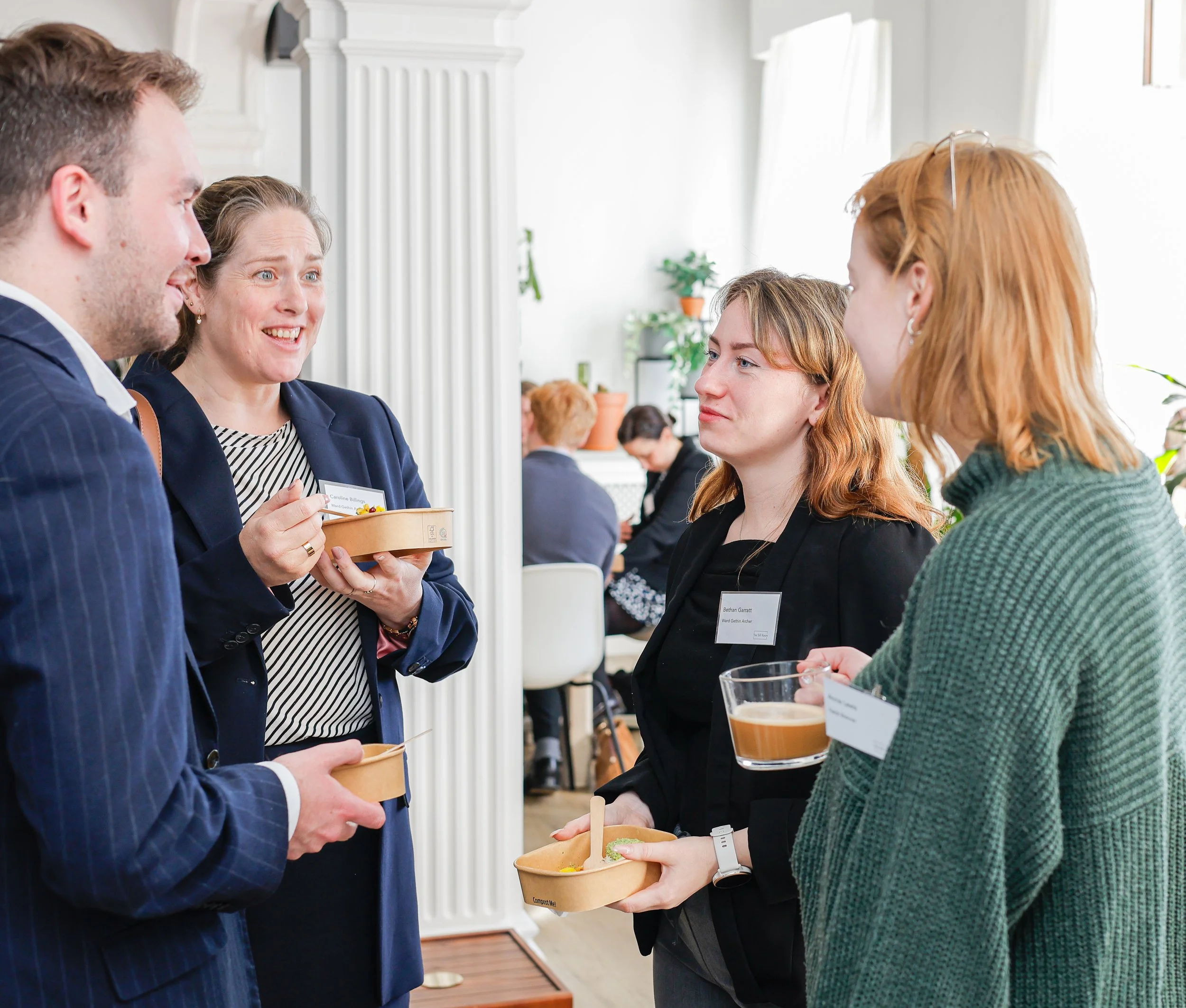 Four people at a social gathering, two women and two men, engaged in conversation while holding small bowls of food and drinks in a brightly lit room with white walls and large windows. The focus is on their interaction, and they are wearing business