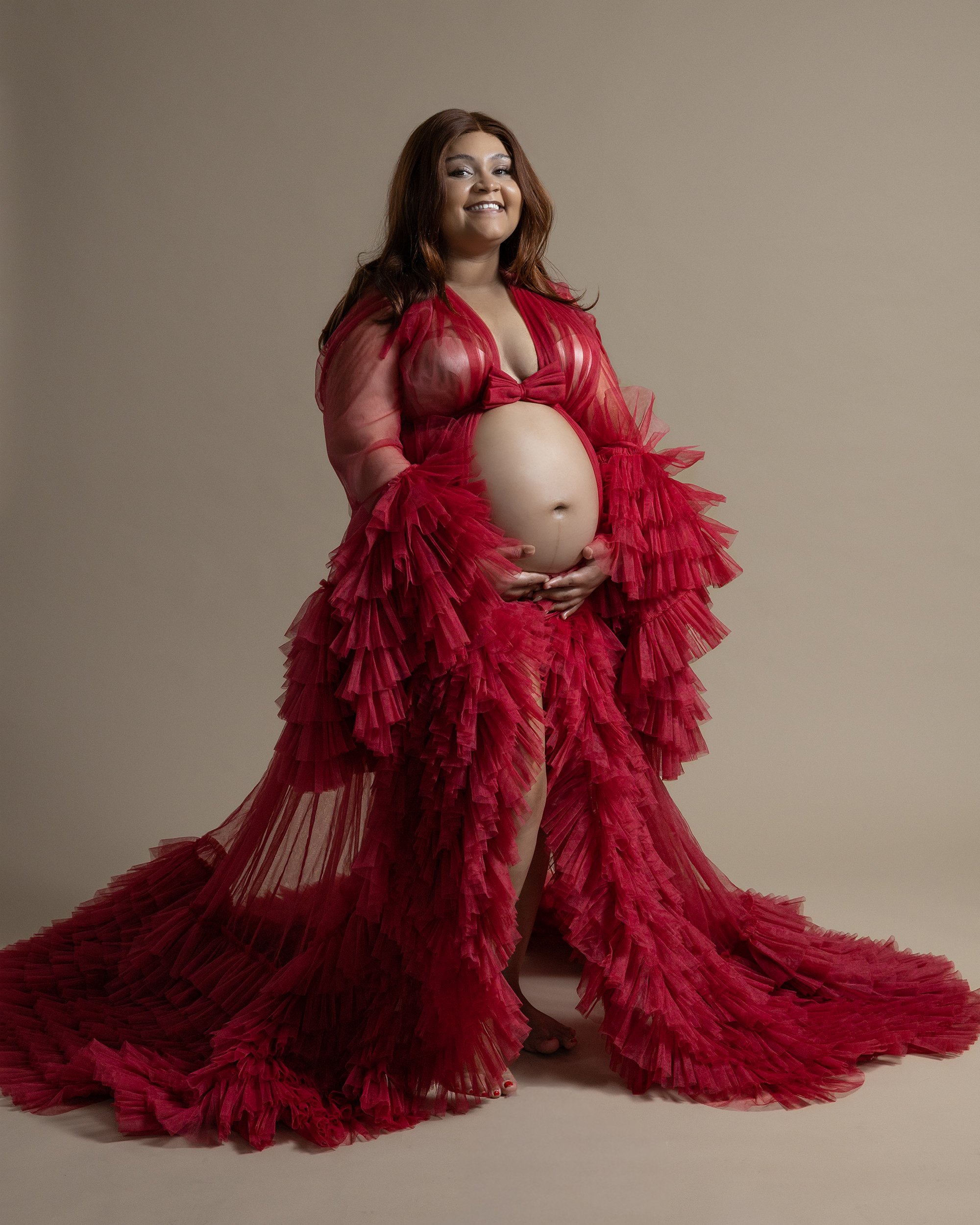 A woman in a flowing red dress with ruffles and sheer fabric, standing against a plain beige background, smiling with hands on her pregnant belly.